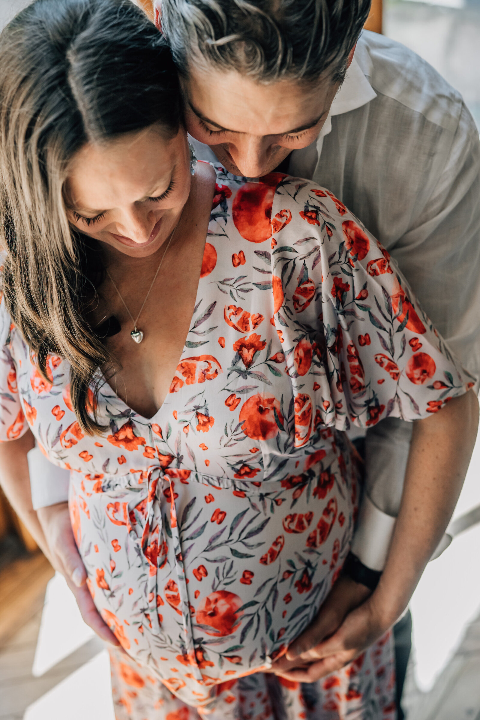 A close-up shot of a pregnant woman in a colorful floral dress, her partner gently embracing her from behind. Both are smiling as they look down at her belly.