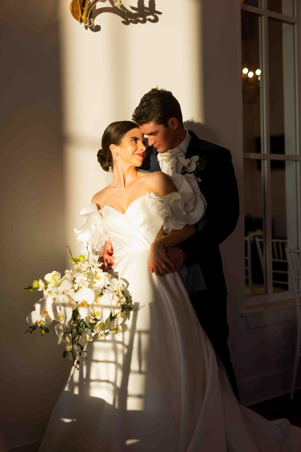 Bride and groom sharing a romantic moment indoors with sunlight highlighting the bride’s gown