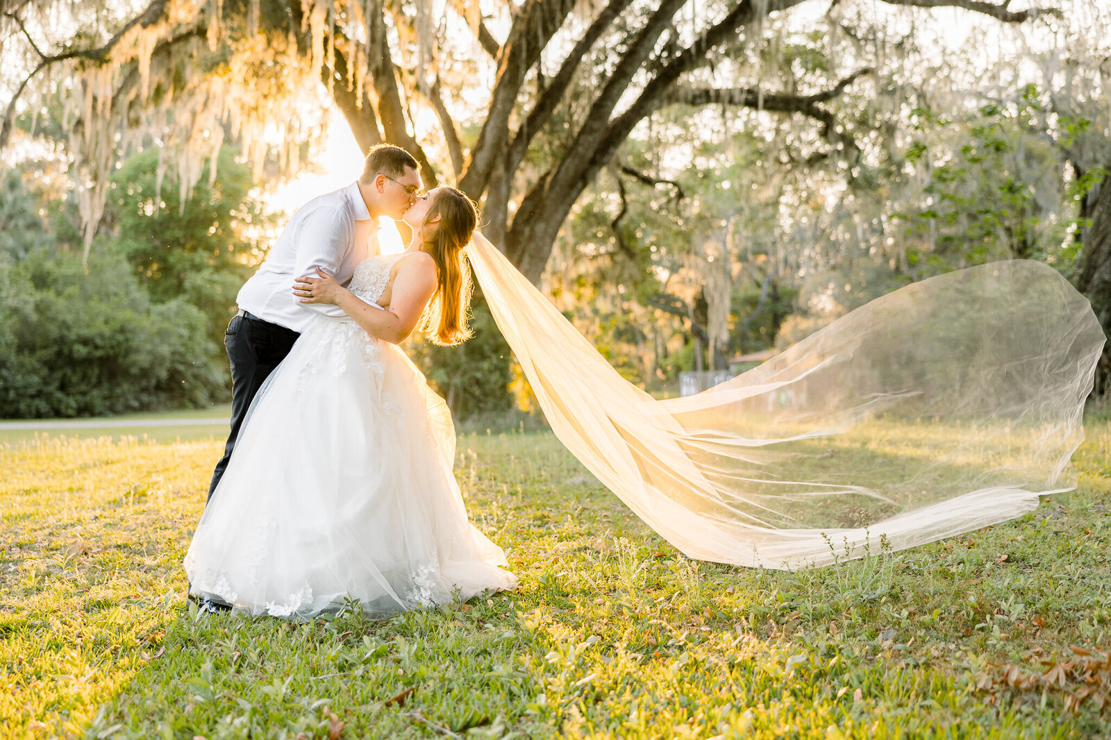 a bride and groom kiss at sunset as her veil floats in the air