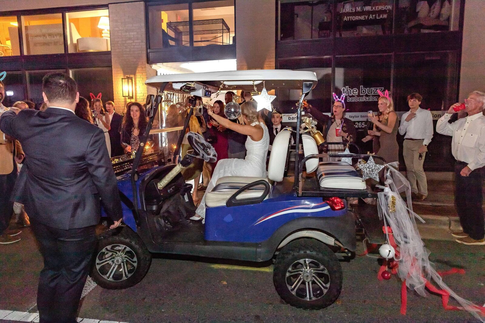 A bride in a white dress sits in a decorated golf cart at night, surrounded by people celebrating. The cart has streamers and cans attached to the back, and the crowd is smiling and taking photos on a city street.