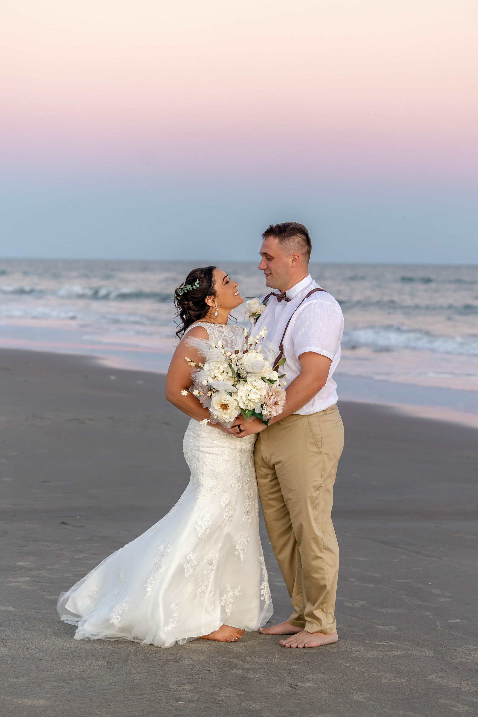 The bride and groom smile at each other barefoot on the beach at sunset, with soft pastel skies in the background, captured during a Ocean Isle, NC wedding, showcasing romance and coastal charm.