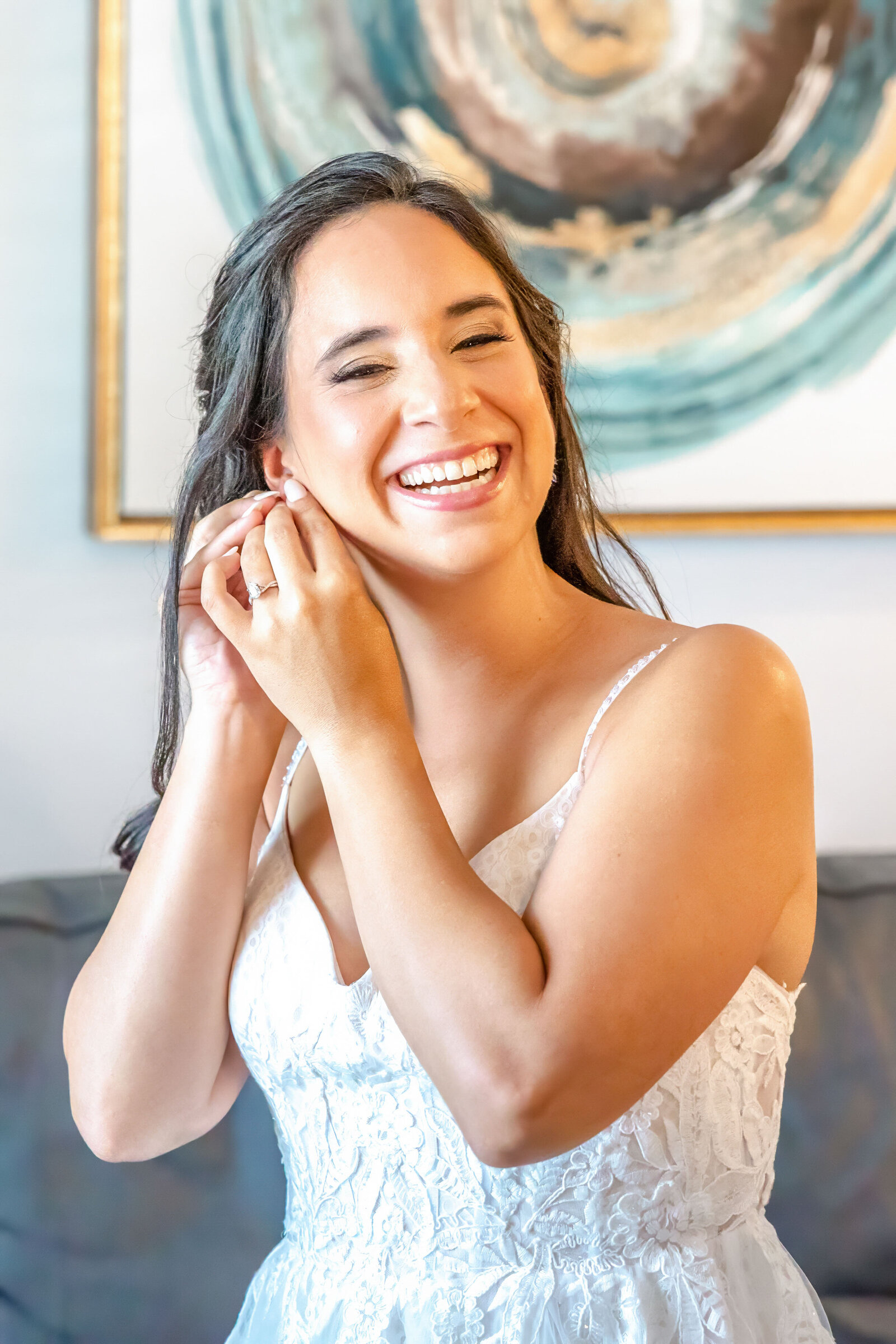 A joyful bride puts on her earrings while smiling in a bright bridal suite, captured during a Wilmington, NC wedding, showcasing authentic preparation moments and timeless elegance.