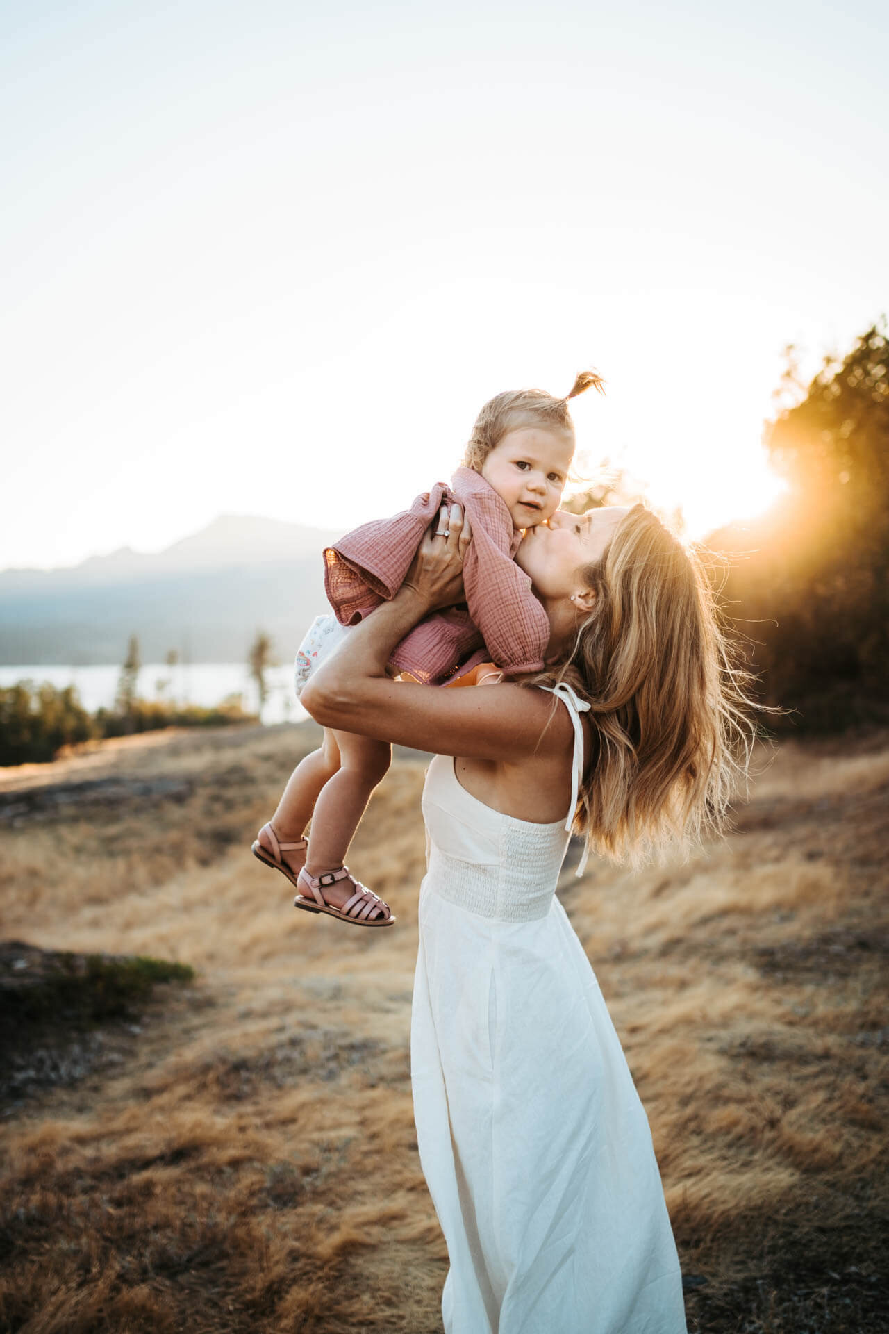 Mom in a white dress holding and  kissing her toddler in golden light