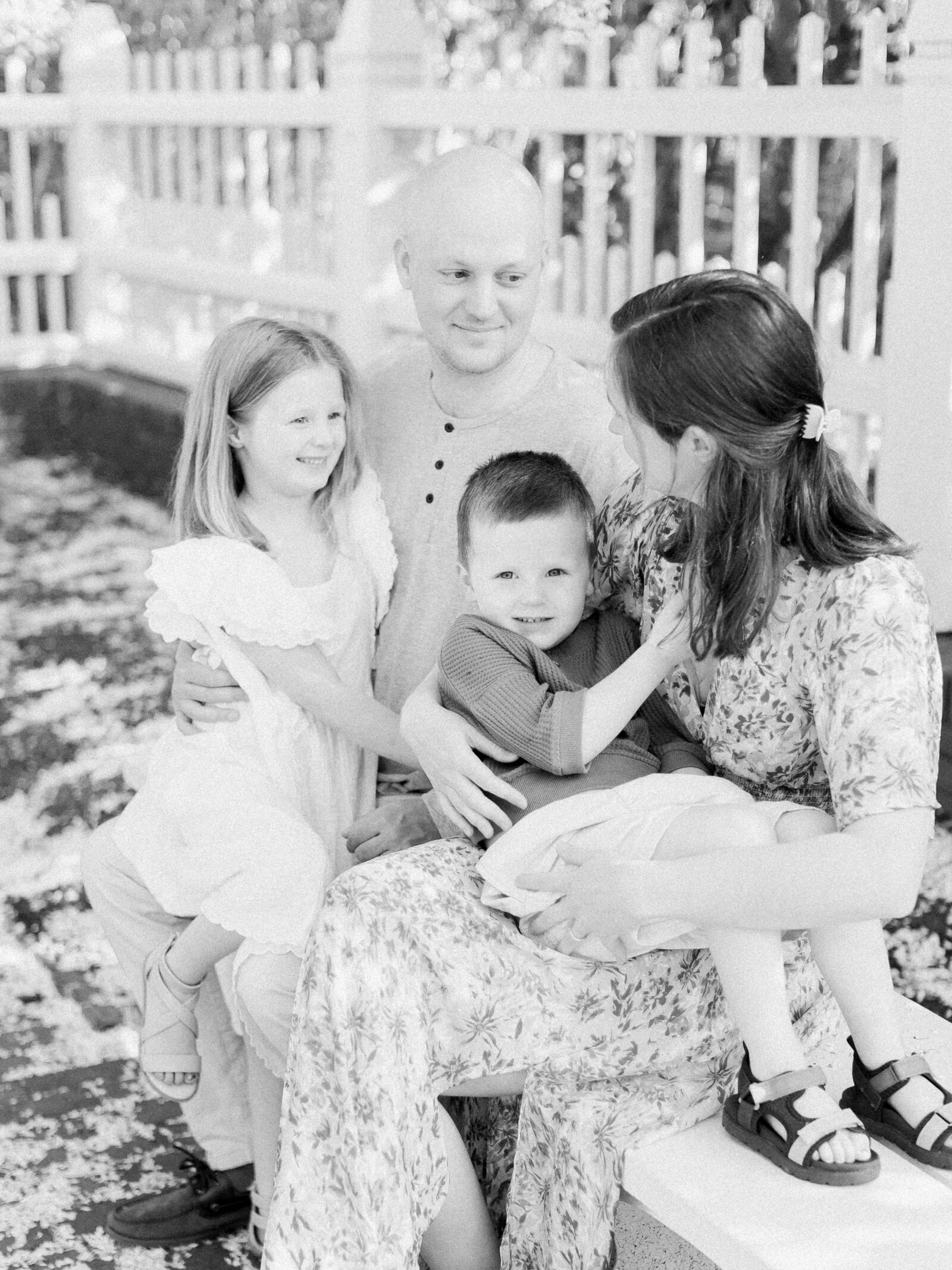 Black and white photo of family of four sitting on a park bench laughing together in a spring garden surrounded by a white picket fence by NH newborn photographer Fieldstone Studio.