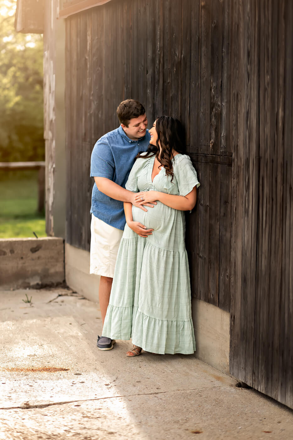 A couple stands close together by a wooden wall. In this tender moment of pregnancy photography, the woman in a light green dress cradles her belly while the man embraces her, both smiling at each other. A Bloomington maternity photographer beautifully captures their joy.