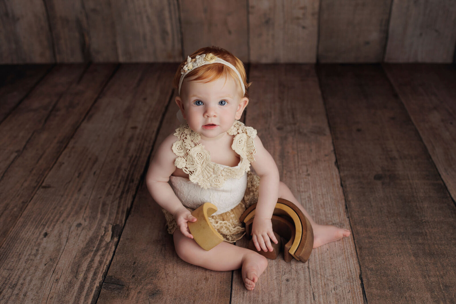 An adorable baby with light hair and blue eyes sits on a rustic wooden floor, wearing a cream-colored lace romper and a matching floral headband. The baby holds a wooden toy, looking up at the camera with a curious expression, creating a charming and vintage-inspired scene.