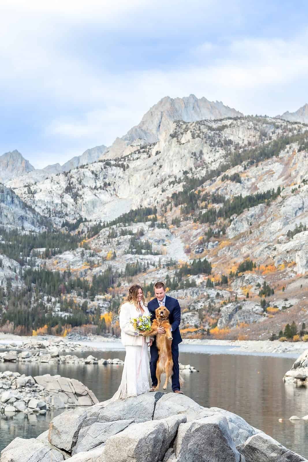 A bride and groom stand in front of a big mountain view and alpine lake while holding their Golden Retriever on their wedding day. 