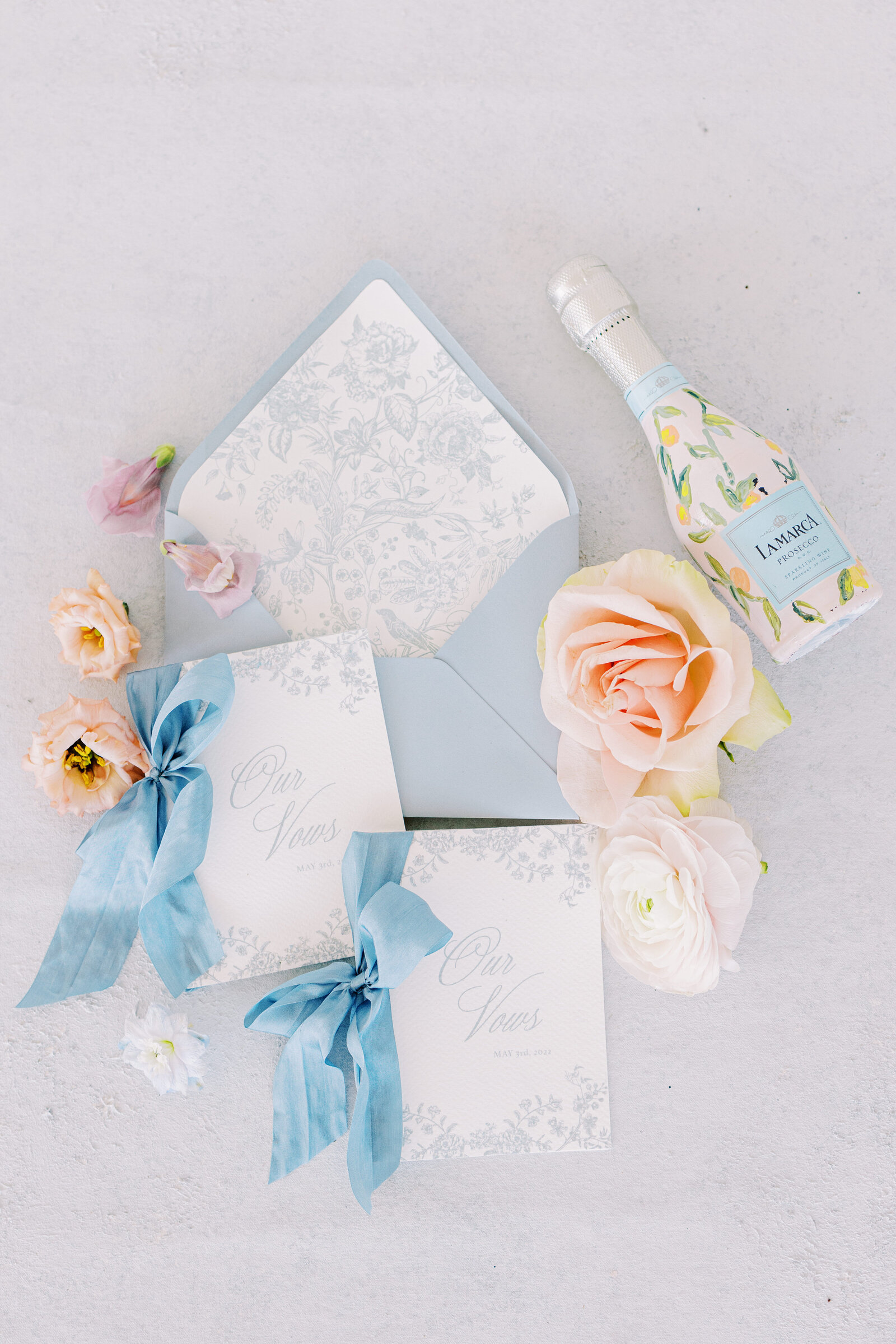 Flat lay details in Charleston, SC, with a blue and white theme for invitations and vow books with some peach and pink flowers and a hand painted champagne bottle
