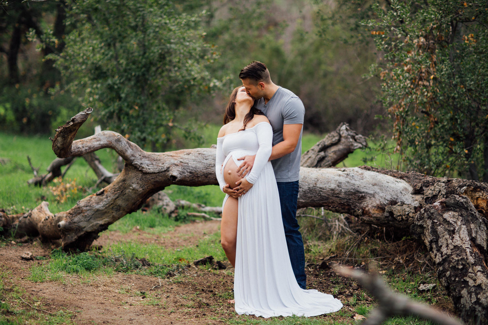 Pregnant wife looks back and kisses her husband as he embraces her during a maternity photo shoot in the woods