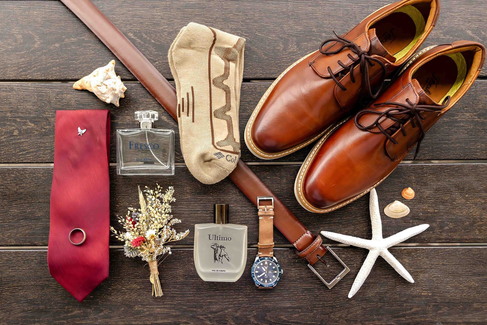 A groom’s flat lay featuring leather shoes, a red tie, a watch, cologne, and coastal accents like seashells and starfish, captured during a Wrightsville Beach, NC wedding, showcasing creative and personal wedding details.