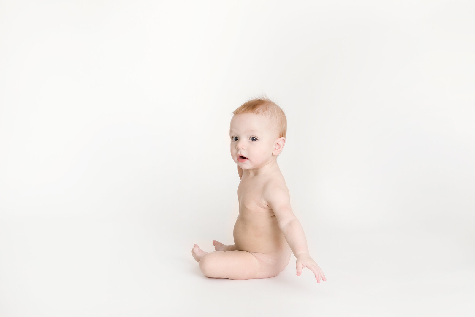 Milestone Photographer , a baby sits up looking curiously on a blank studio backdrop