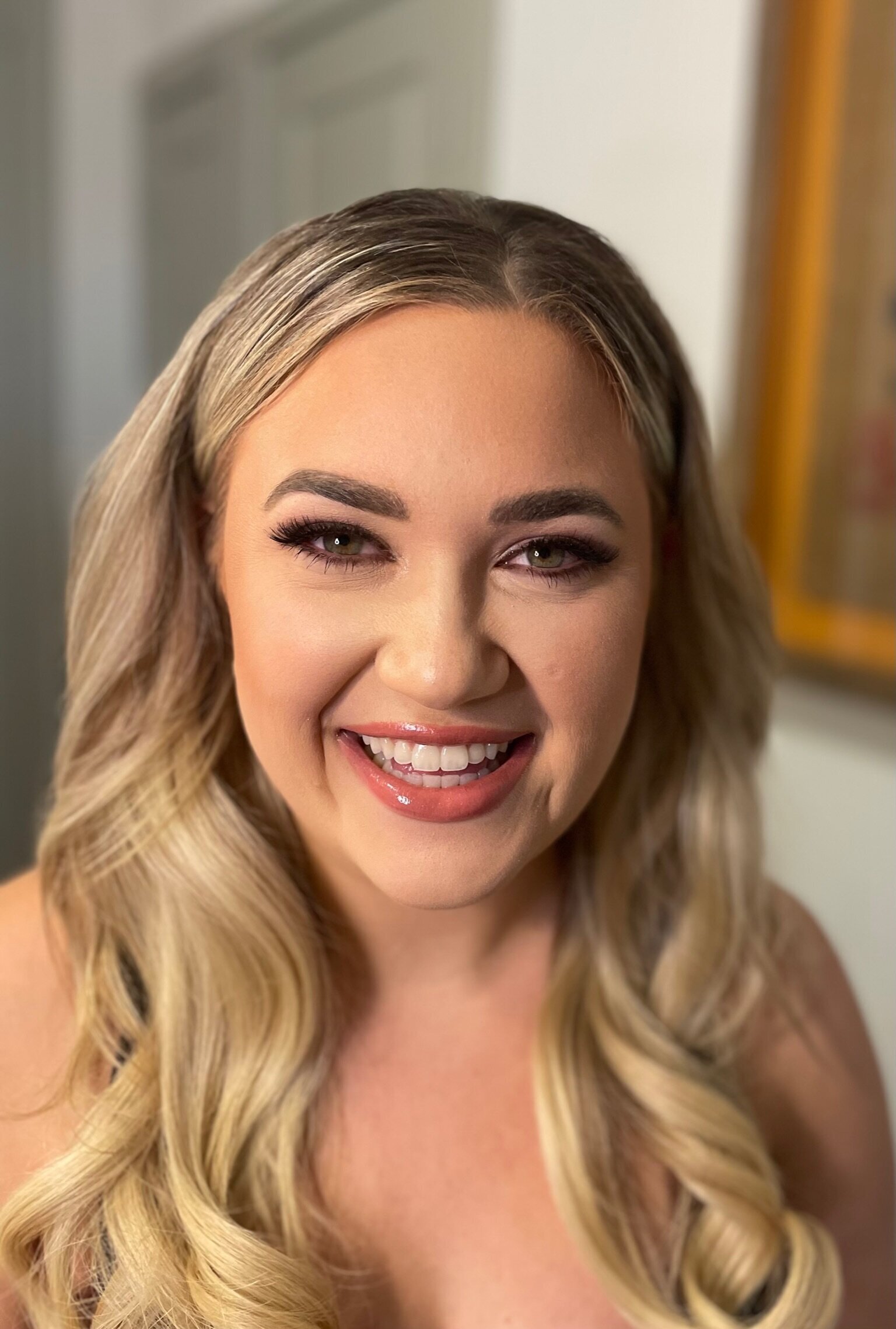 A woman with long blonde hair smiles in a close-up shot, showcasing her radiant bridal beauty experience. She's indoors, elegantly wearing makeup.