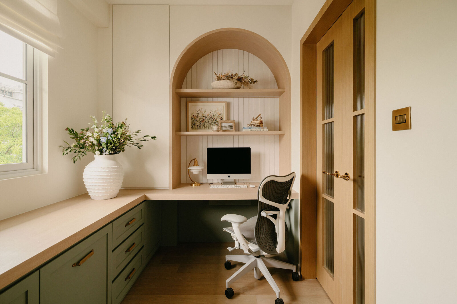 Home office nook with arched shelving, built-in timber desk, and contemporary ergonomic chair framed by natural light.