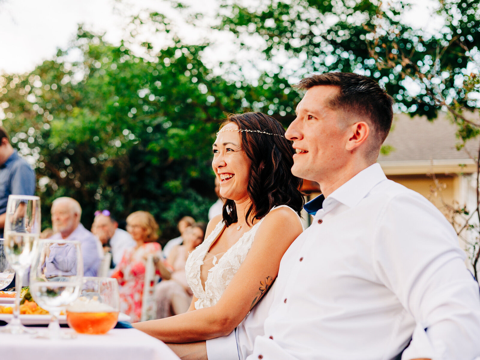 A smiling couple in formal attire sits at a table outdoors, enjoying an event with other guests in the background. Captured by an Eau Claire Wedding Photographer, the scene is bright and cheerful with green trees and soft sunlight.