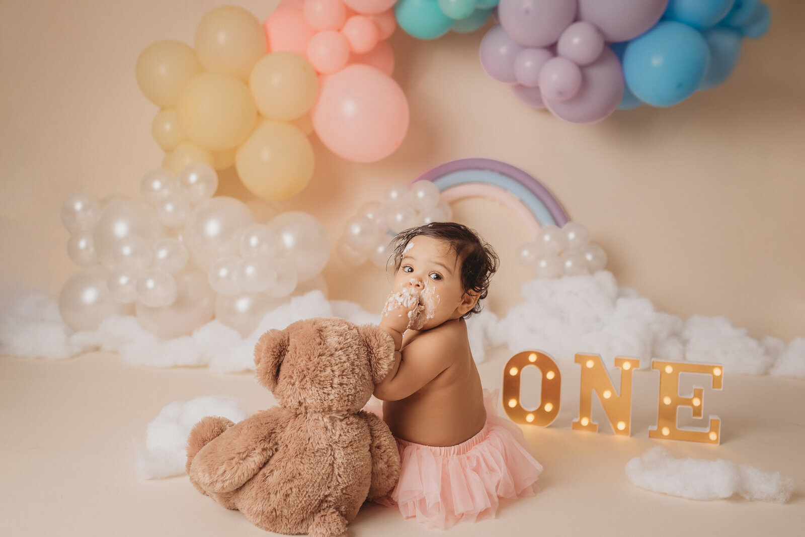 one year old baby eating a smash cake at Milton GA cake smash session sitting on cream backdrop with pastel rainbow balloon garland in background
