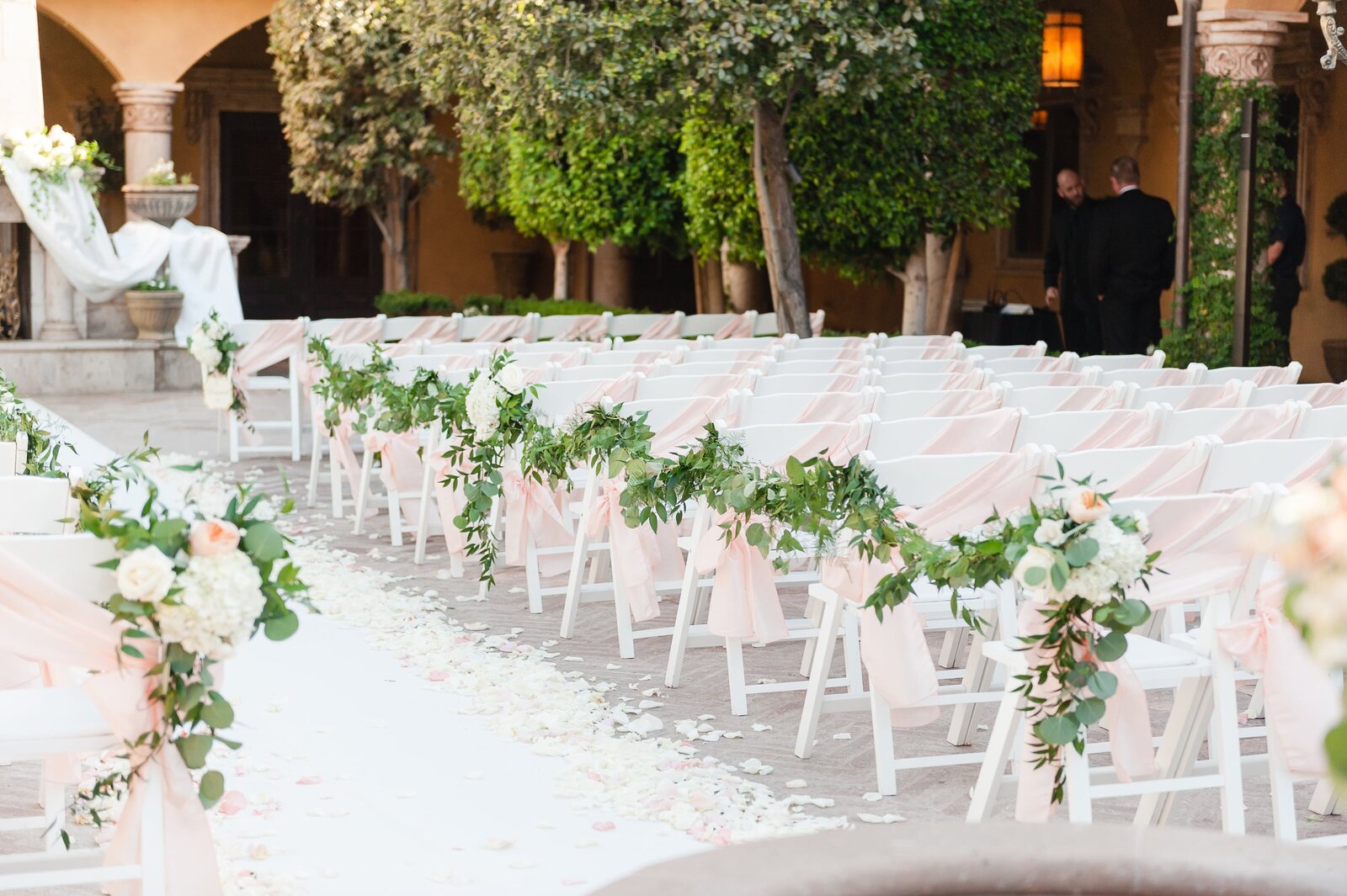 Villa Siena ceremony setting with white chairs draped with green swag and blush sashes.