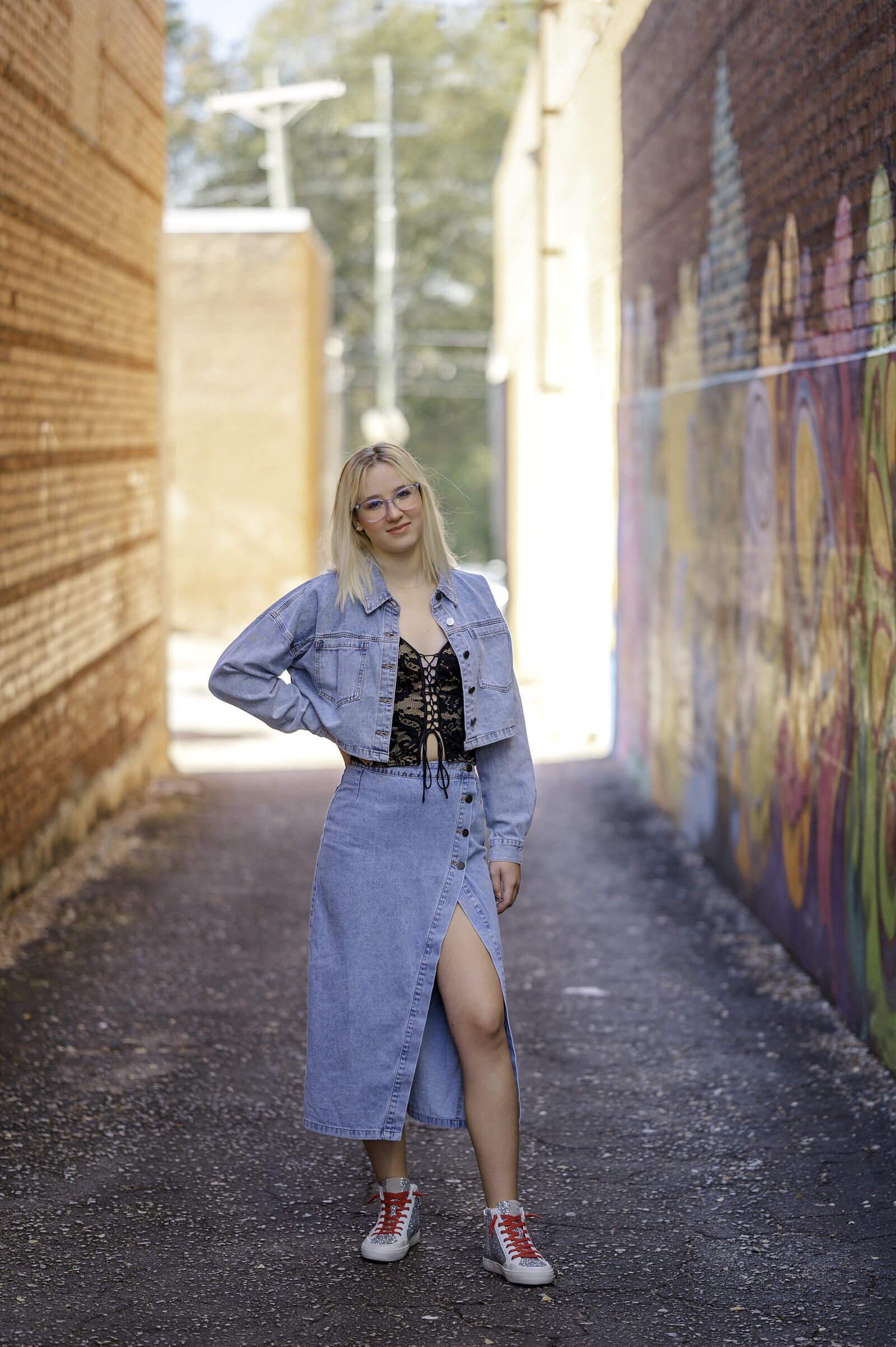 Beautiful blonde senior stands between two buildings with her hand on her hip and smiles