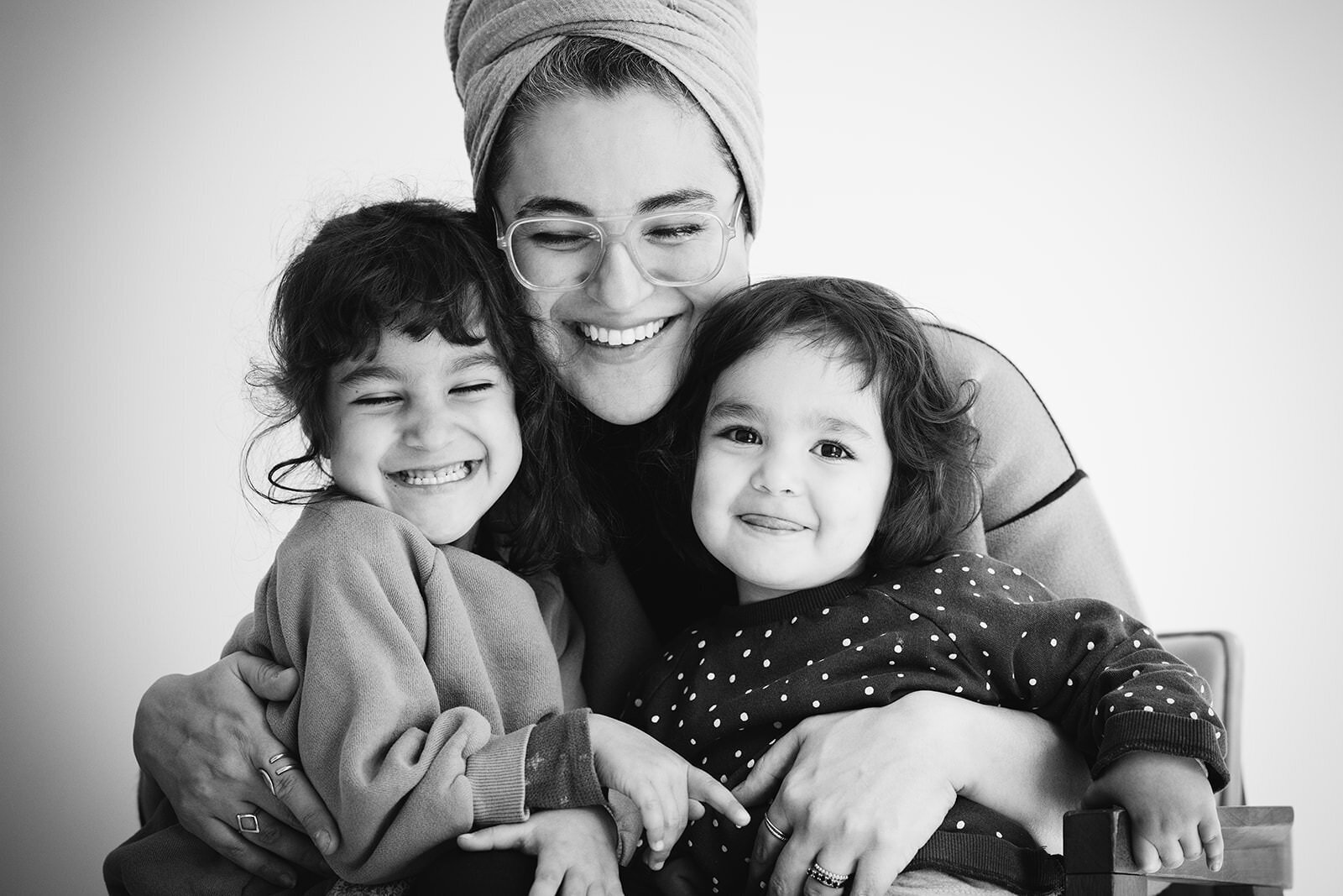 A joyful black-and-white portrait of a mother wearing glasses and a headscarf, smiling warmly while embracing her two young daughters, radiating love and connection.
