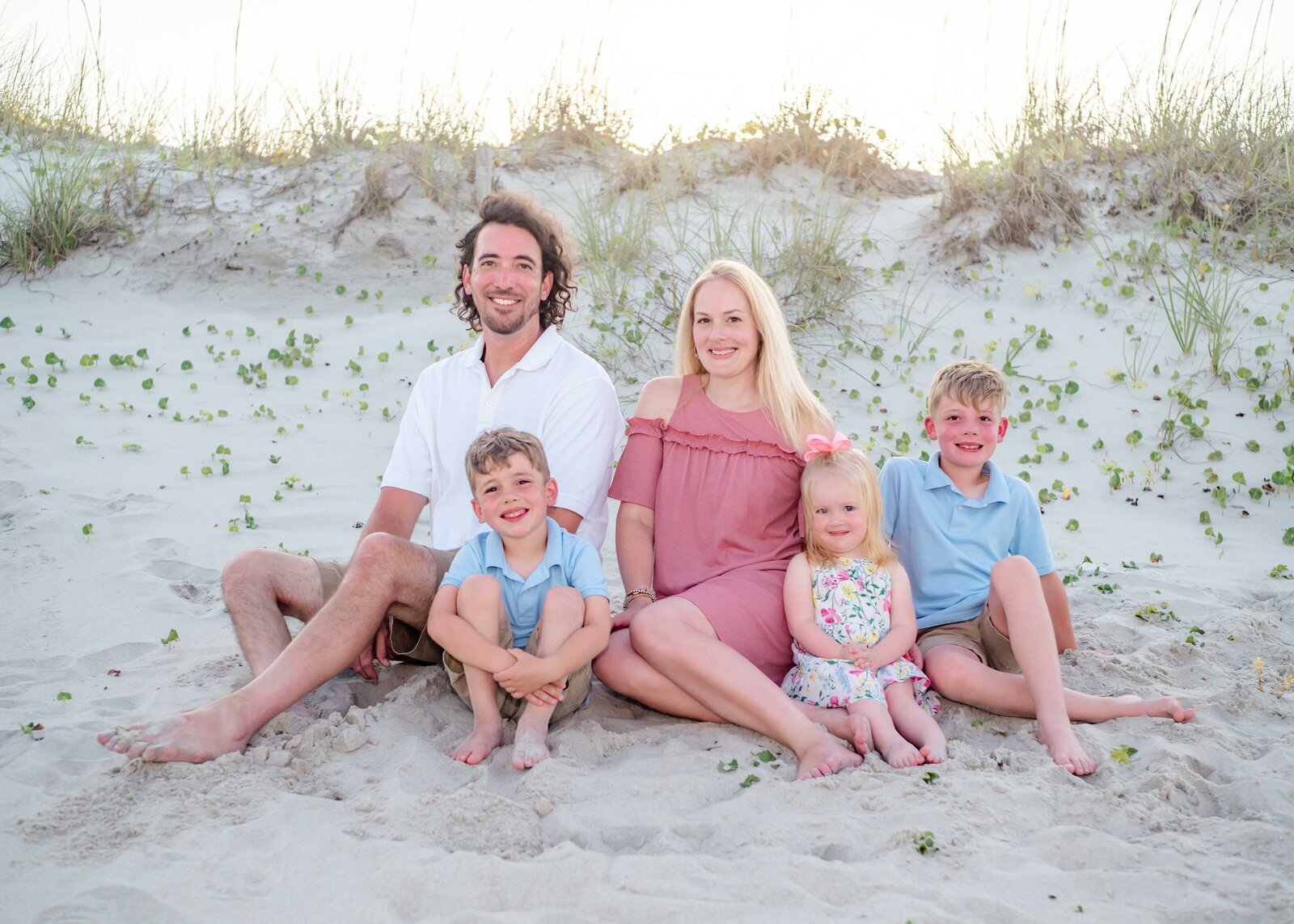 A family of five sits together on sandy dunes at sunset, dressed in pastel colors. The parents and children share warm smiles, surrounded by beach grass. This image showcases the best family photographer in North Carolina, perfect for capturing intimate family portraits.