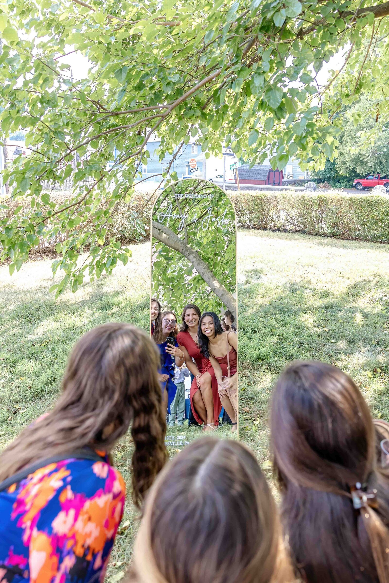 Bridesmaids gather around and smile into a tall mirror placed outdoors under a tree, captured during a Wrightsville Beach, NC wedding, highlighting fun and creative moments.