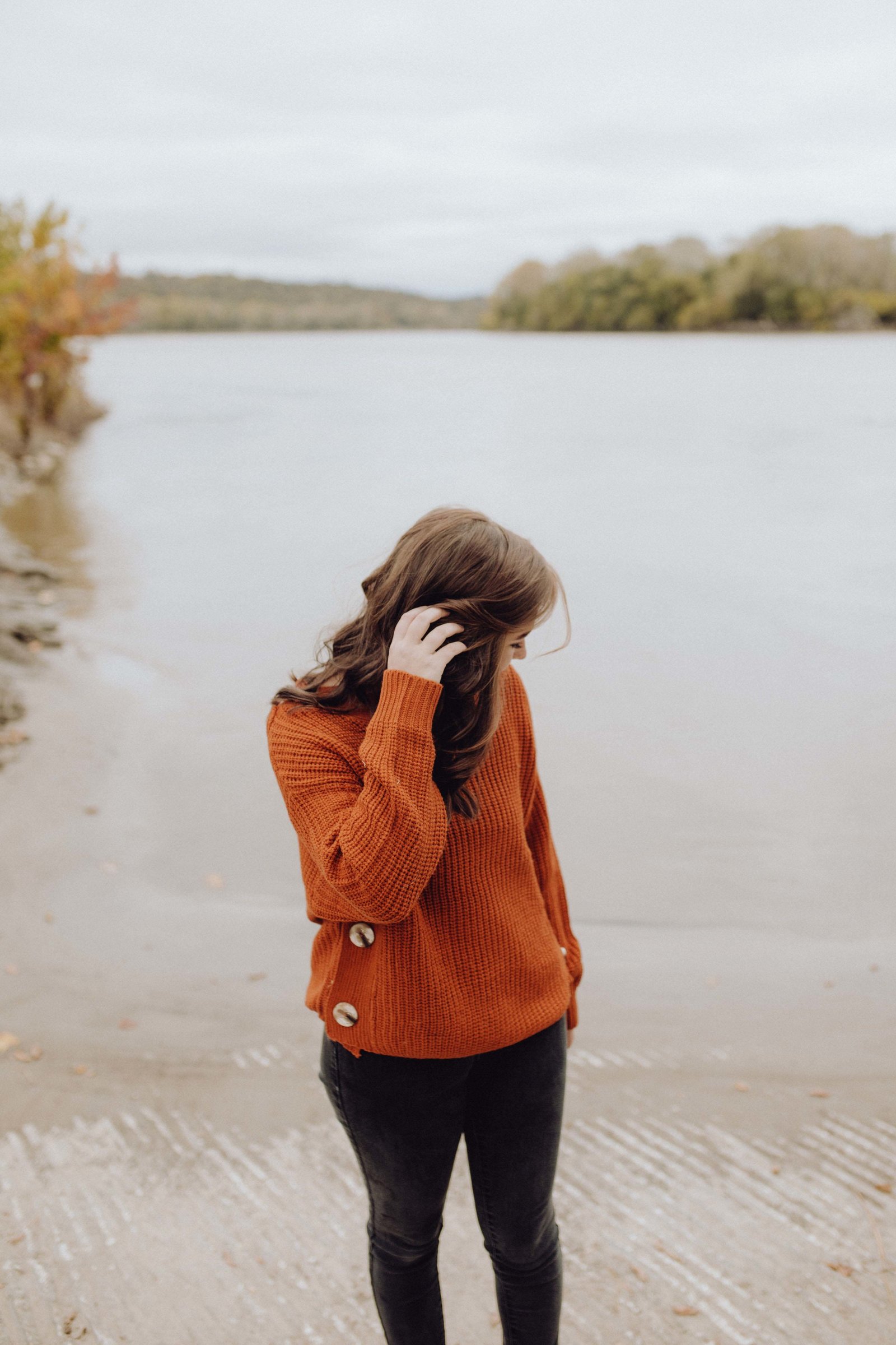 Girl is standing by river water, hand in her hair and looking at water.