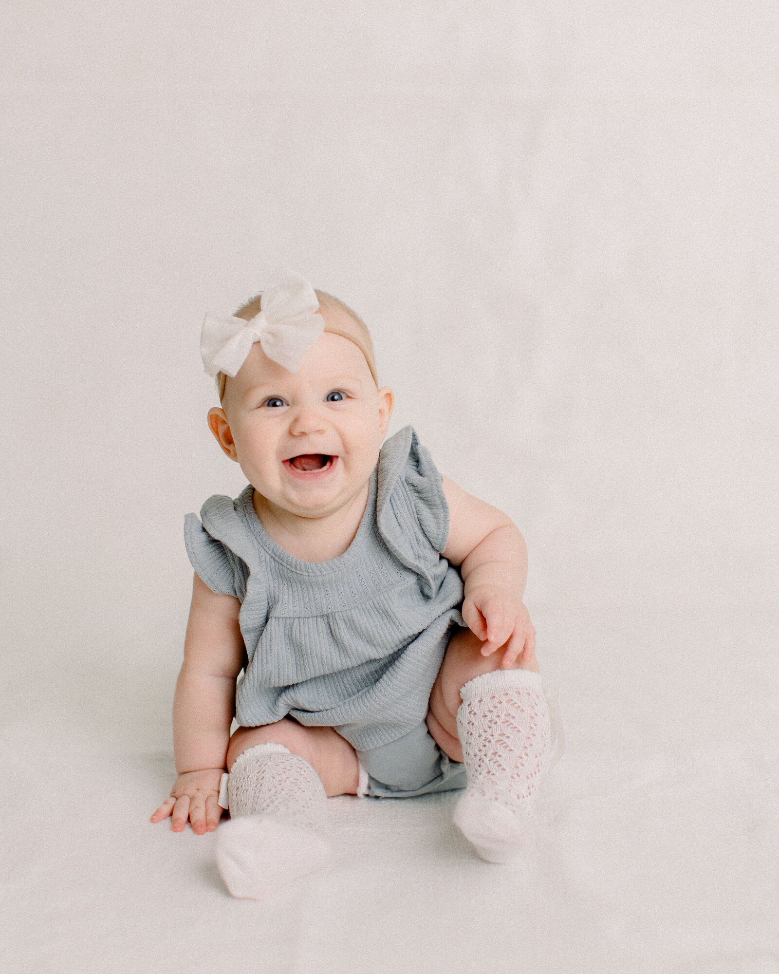 Happy laughing baby sitting up in a blue romper and white bow in a neutral studio by NH newborn photographer Fieldstone Studio.