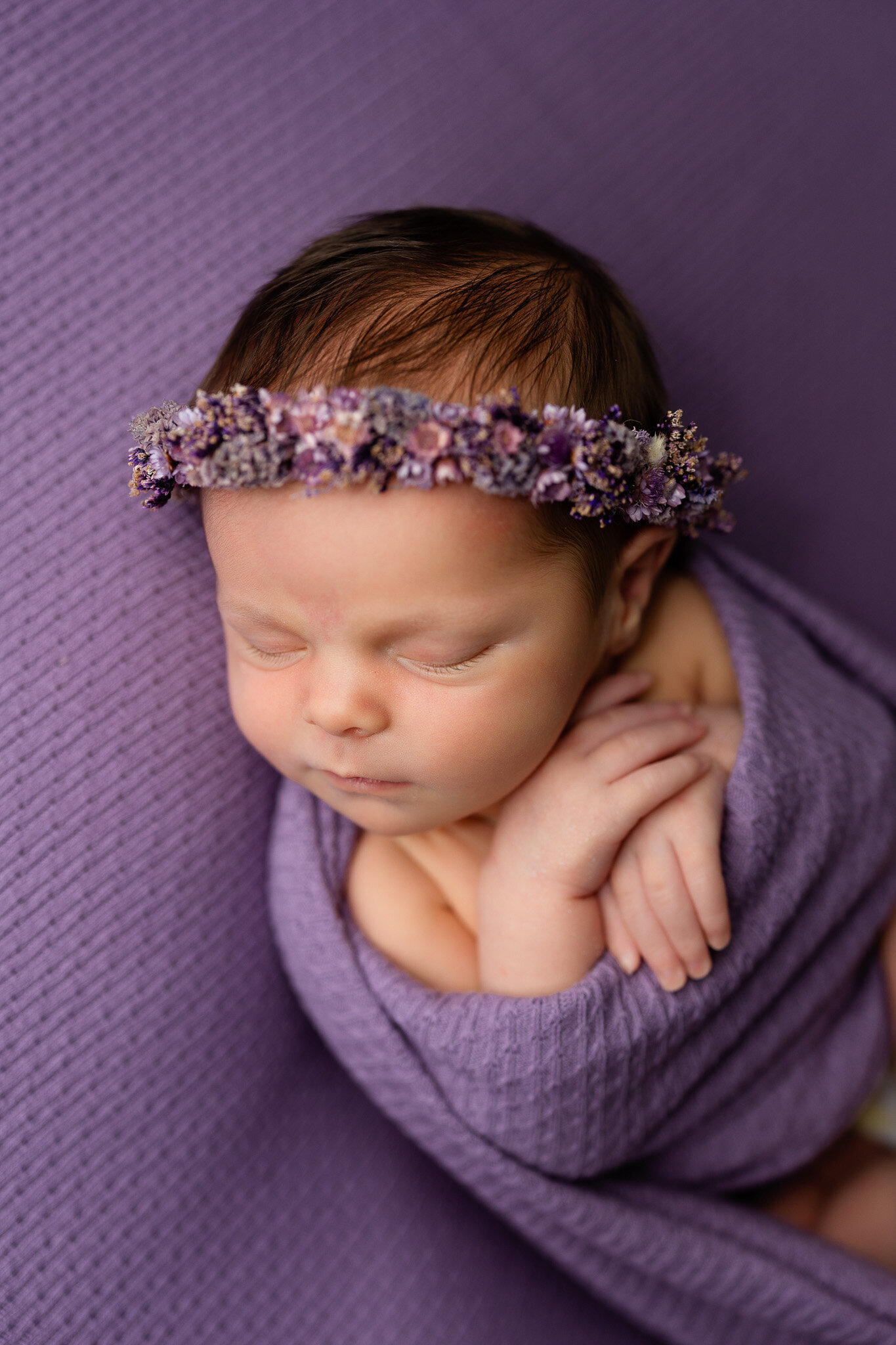 Baby girl wrapped in lavender wearing a purple floral headband, posed on a matching back drop