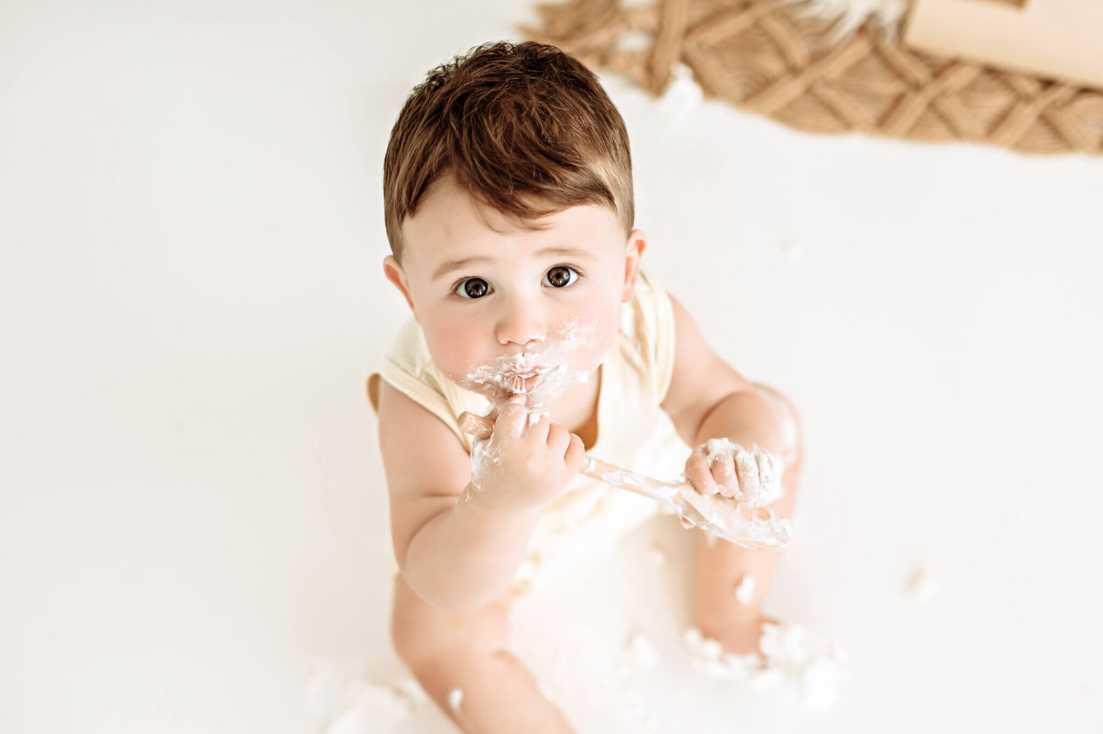 A baby sits on the floor, eating cake with a wooden spoon. The scene is decorated with green, gold, and white balloons in the background. The baby, wearing a light-colored outfit, is playfully covered in cake and frosting.