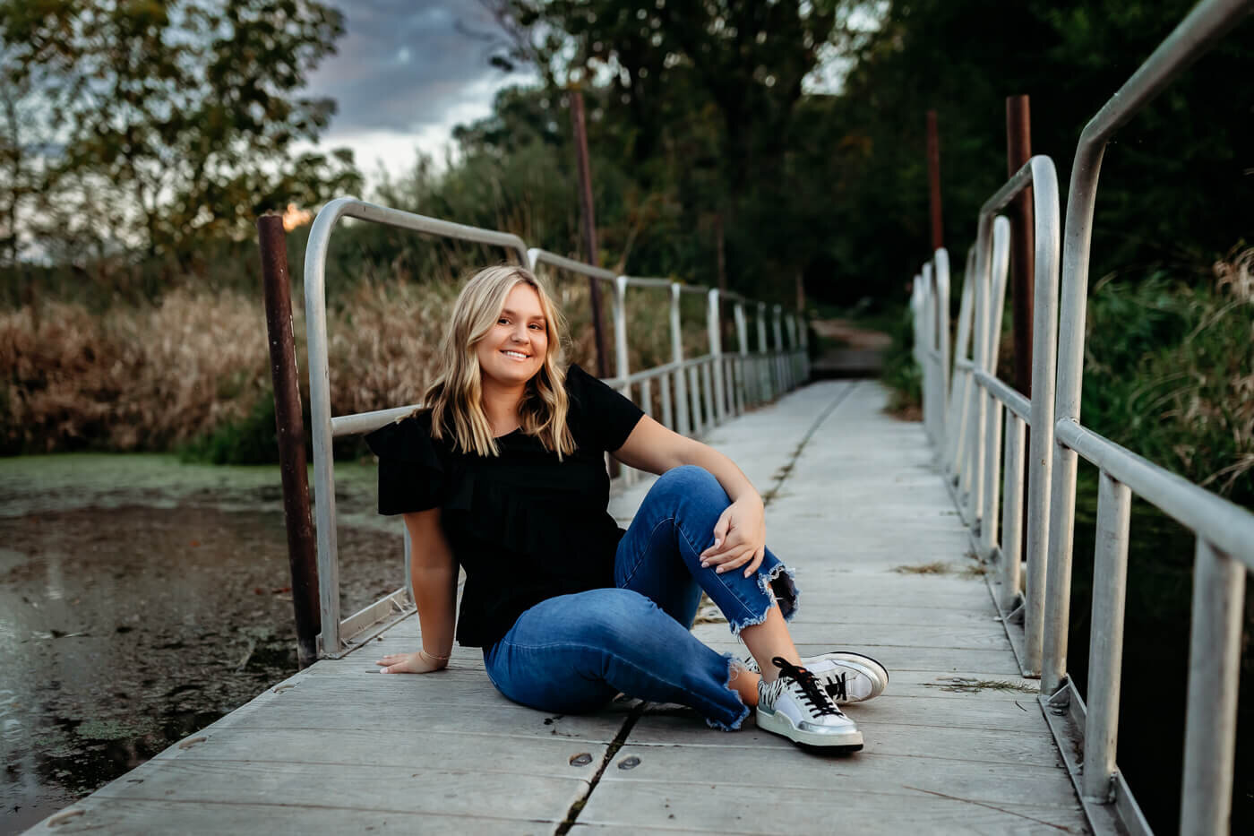 A woman with blonde hair sits on a wooden walkway surrounded by nature. She's wearing a black shirt, blue jeans, and sneakers, smiling at the camera. Trees and grass are visible in the background.