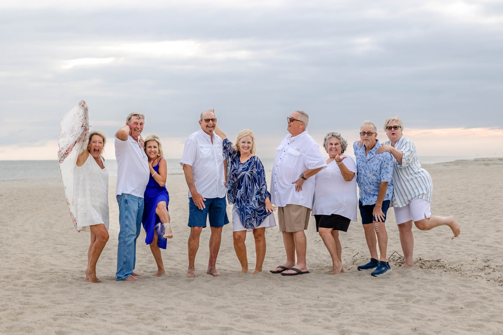 A lively group poses playfully on a cloudy beach, dressed in casual white and blue outfits. Their cheerful expressions capture a fun and relaxed atmosphere, perfect for North Carolina family photography, highlighting joyful beachside gatherings.