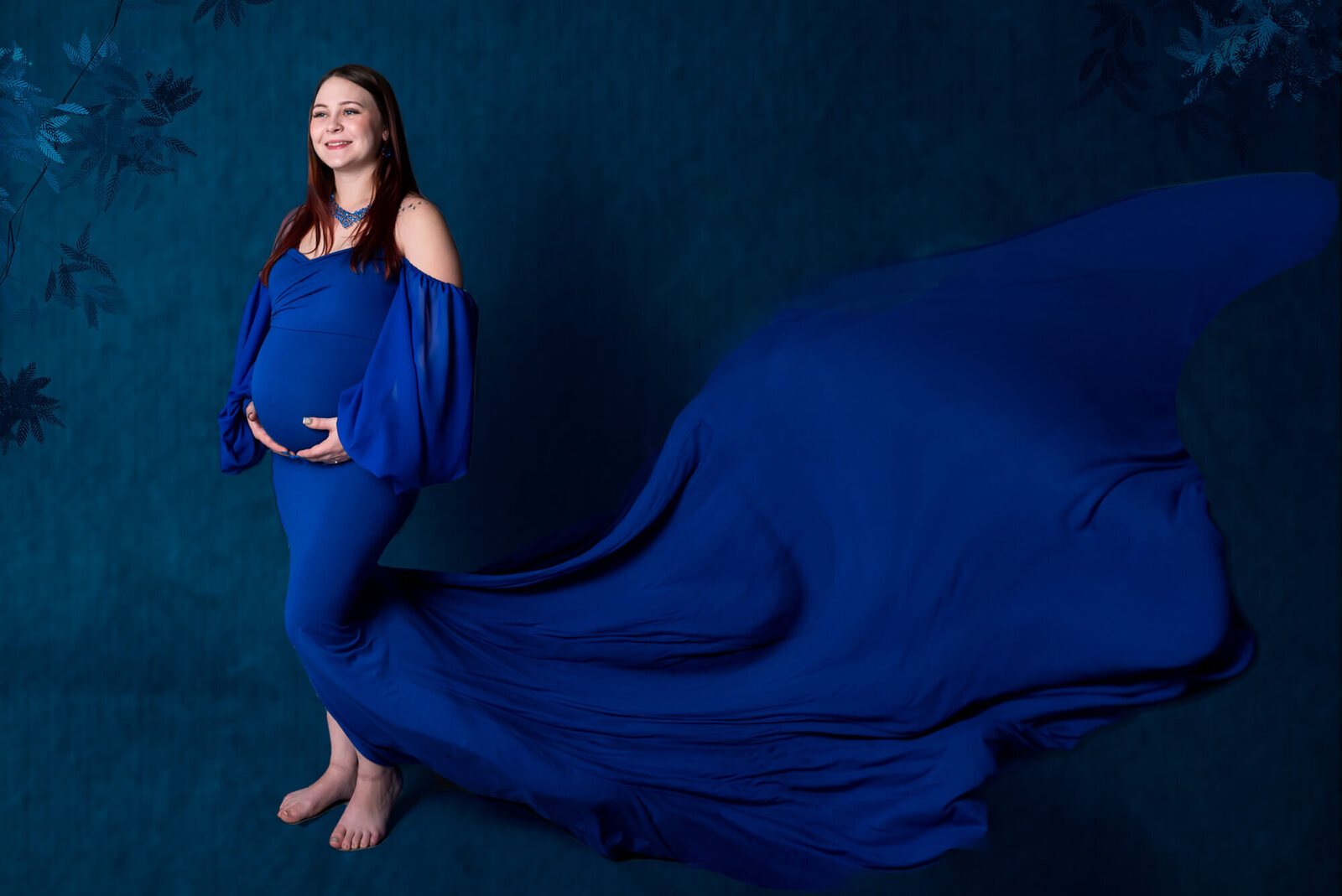   mother-to-be in a flowing royal blue gown stands against a dark blue floral backdrop, her dress trailing behind her for a dramatic effect.