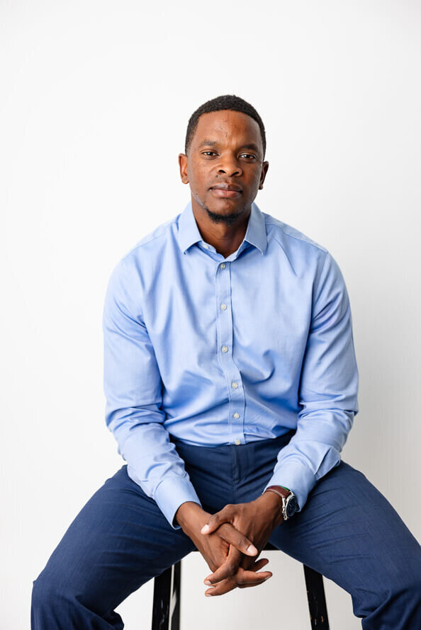 Man with blue shirt in a white studio sits and folds hand in lap for his headshot.