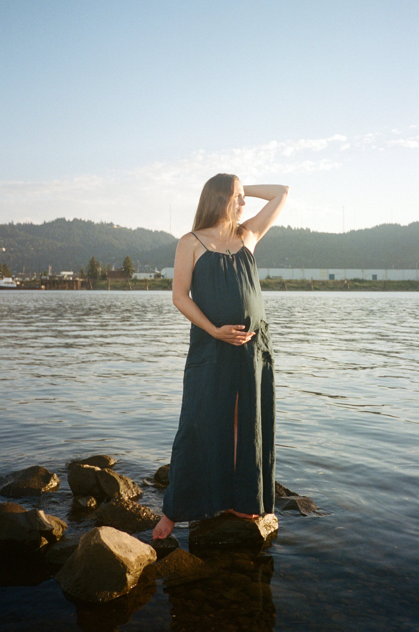 Mother embracing pregnant belly and standing on rock in the Williamette River in Portland.