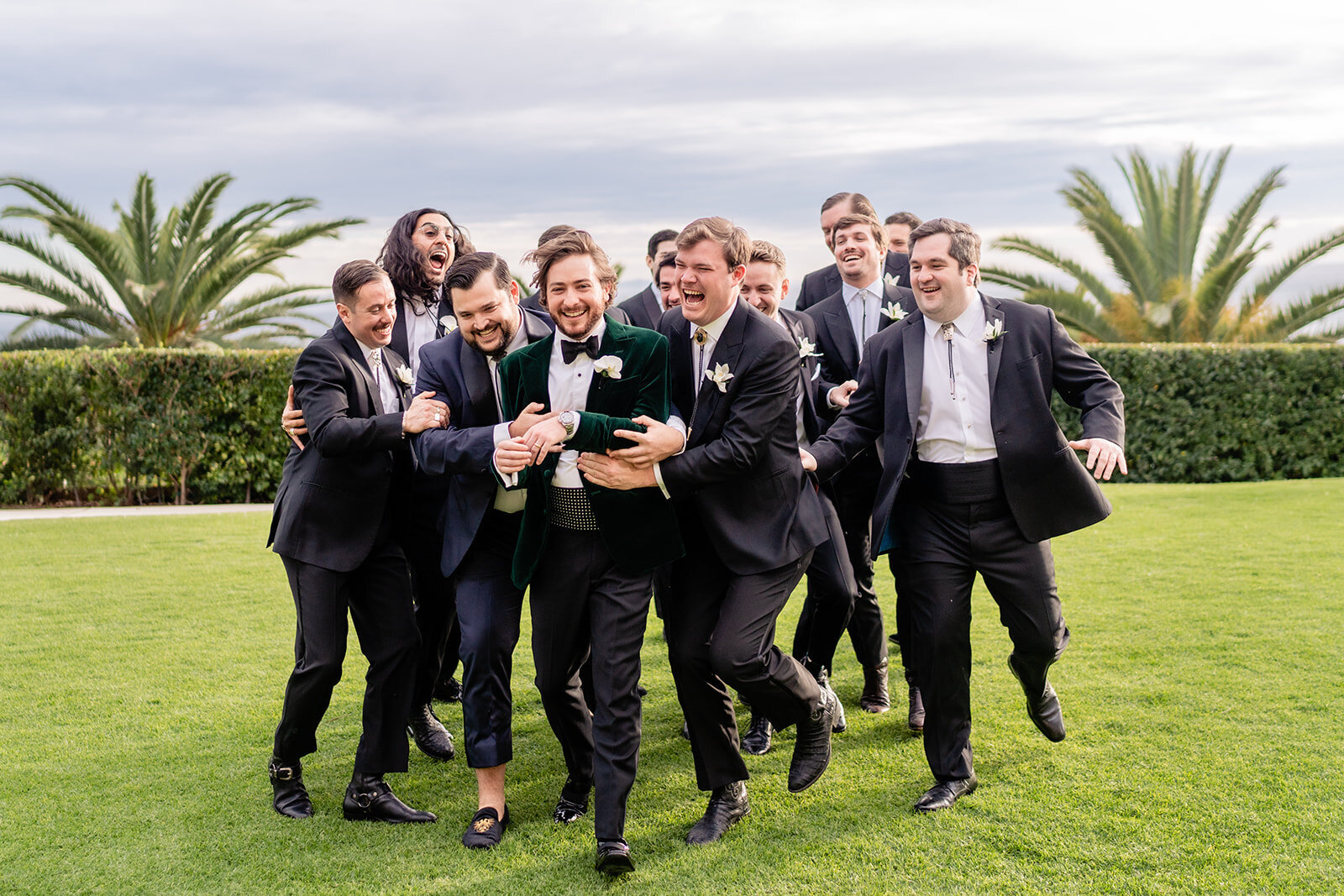 Groomsmen huddled and embracing groom as they are walking on a green lawn with palm trees in the background