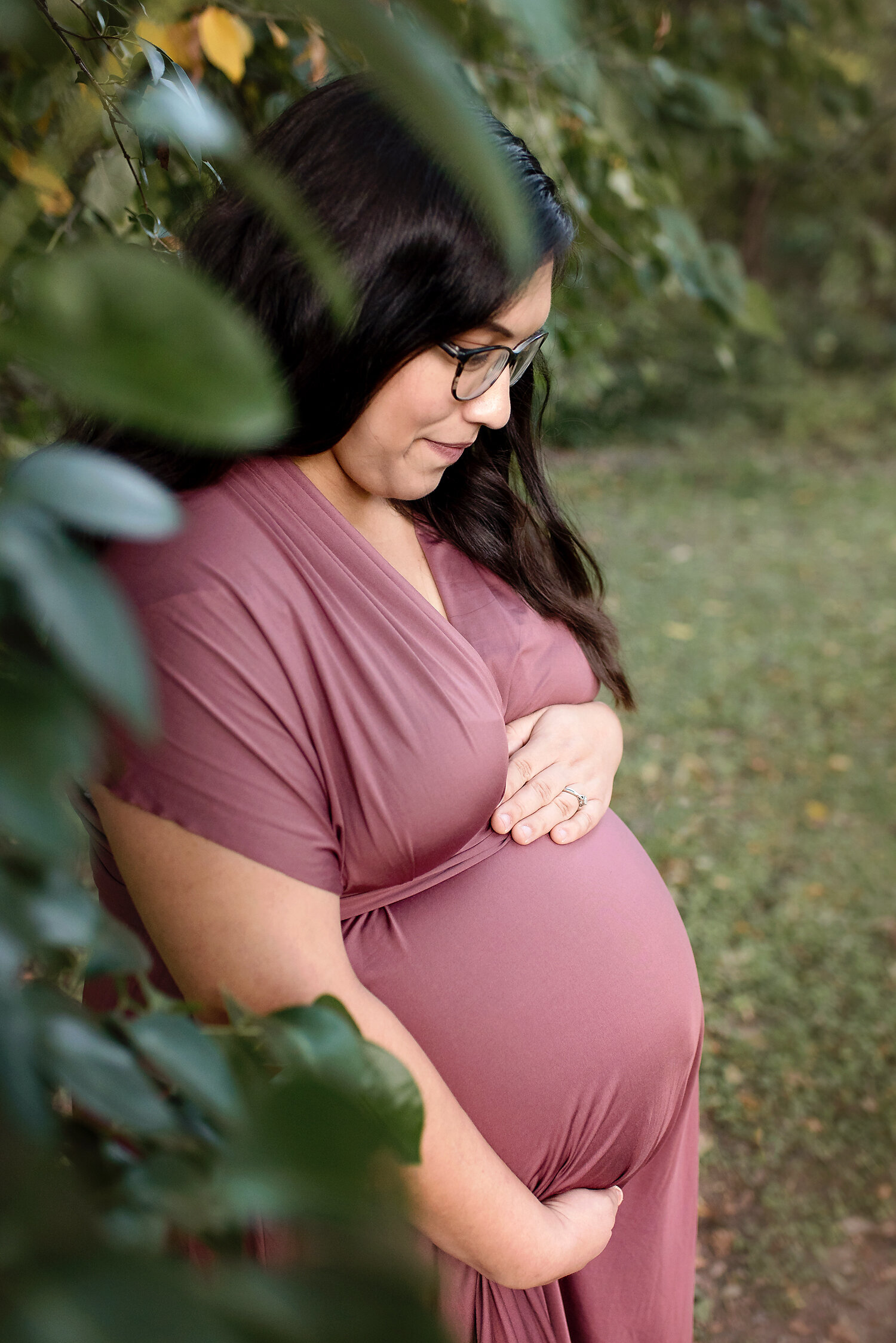 A pregnant woman holding her belly while standing behind leaves.