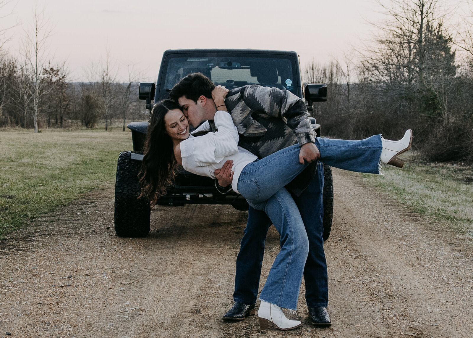 Man holds woman in a dip in front of a black jeep, sharing a kiss. The sun sets colorfully over Shelby Farms Park.