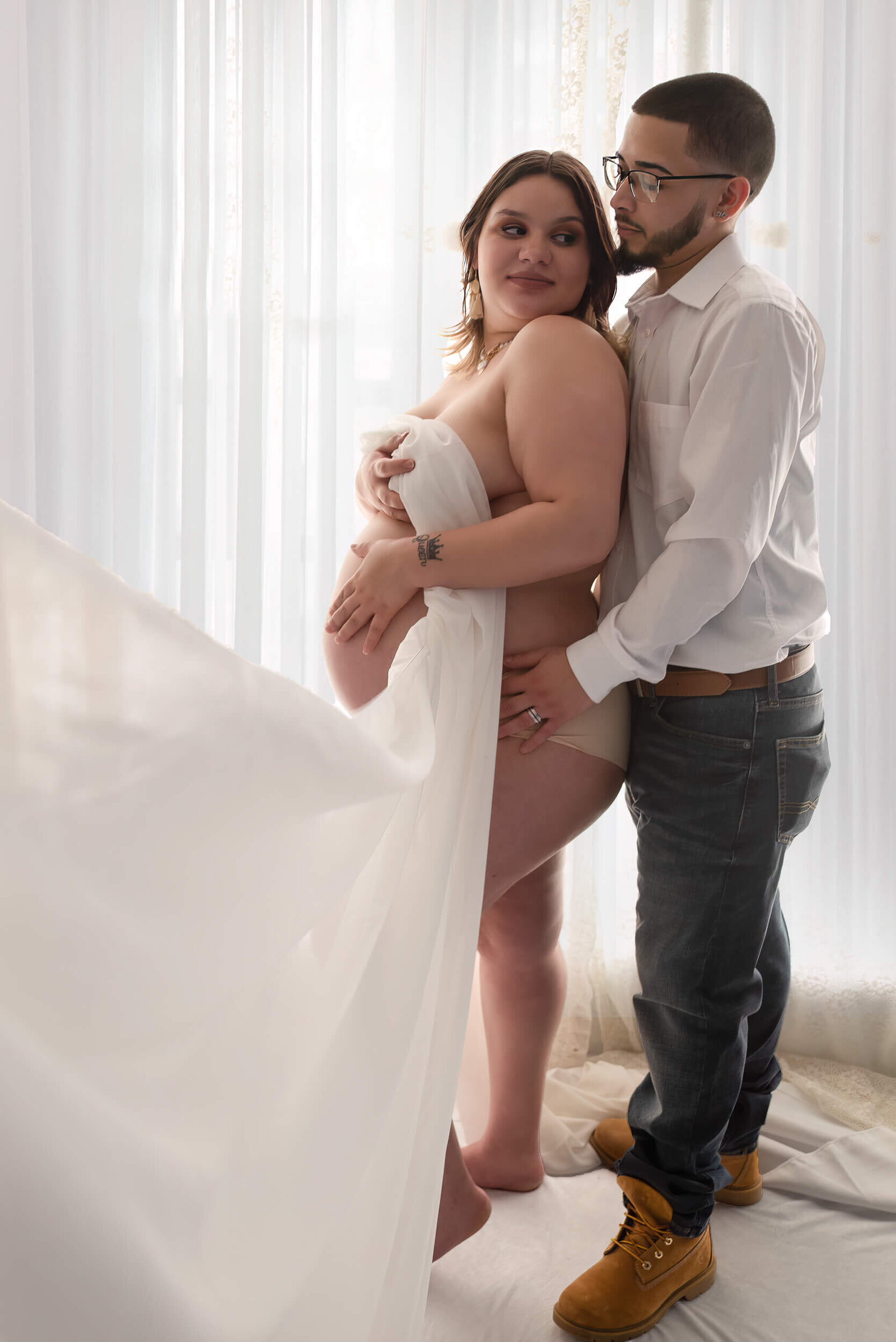A pregnant woman draped in soft white fabric gazes lovingly at her partner, who holds her gently, against a sheer white curtain backdrop.
