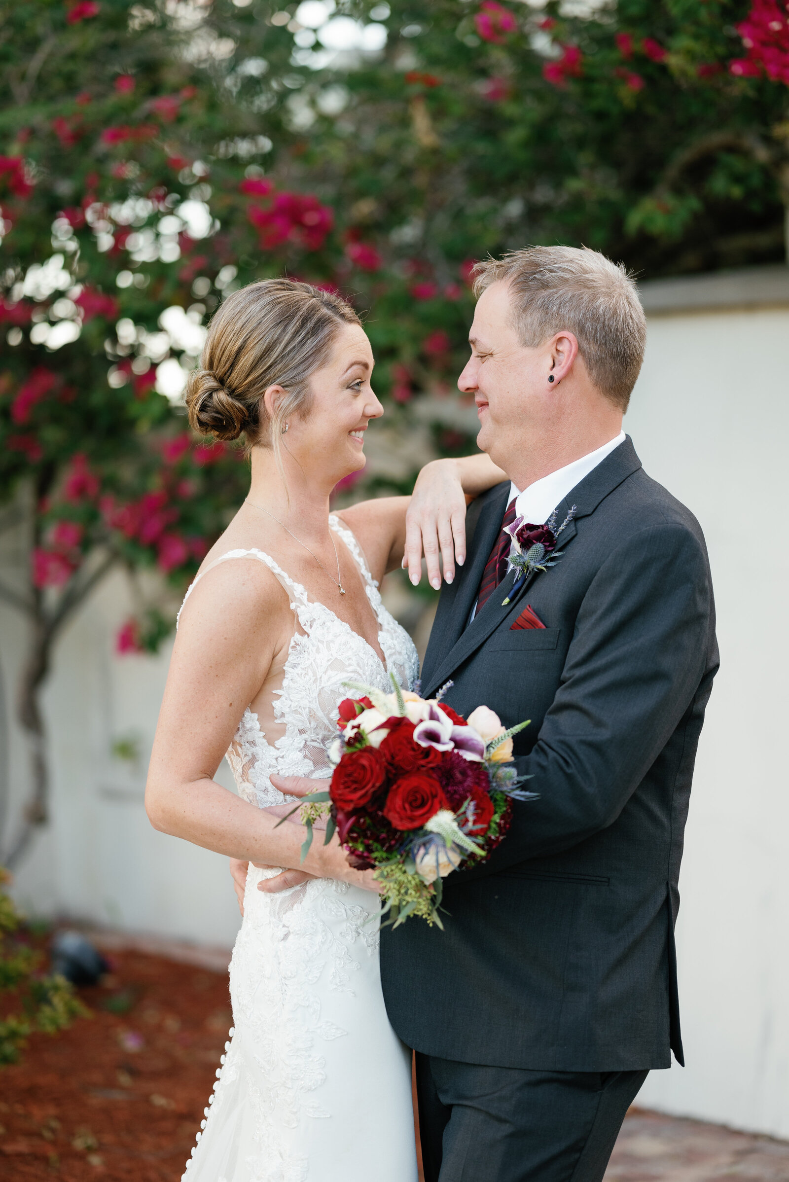 Bride and groom facing each other and smiling. Her left arm is resting on his shoulder and she is holding her wedding bouquet in her right hand. His hands are on her waist