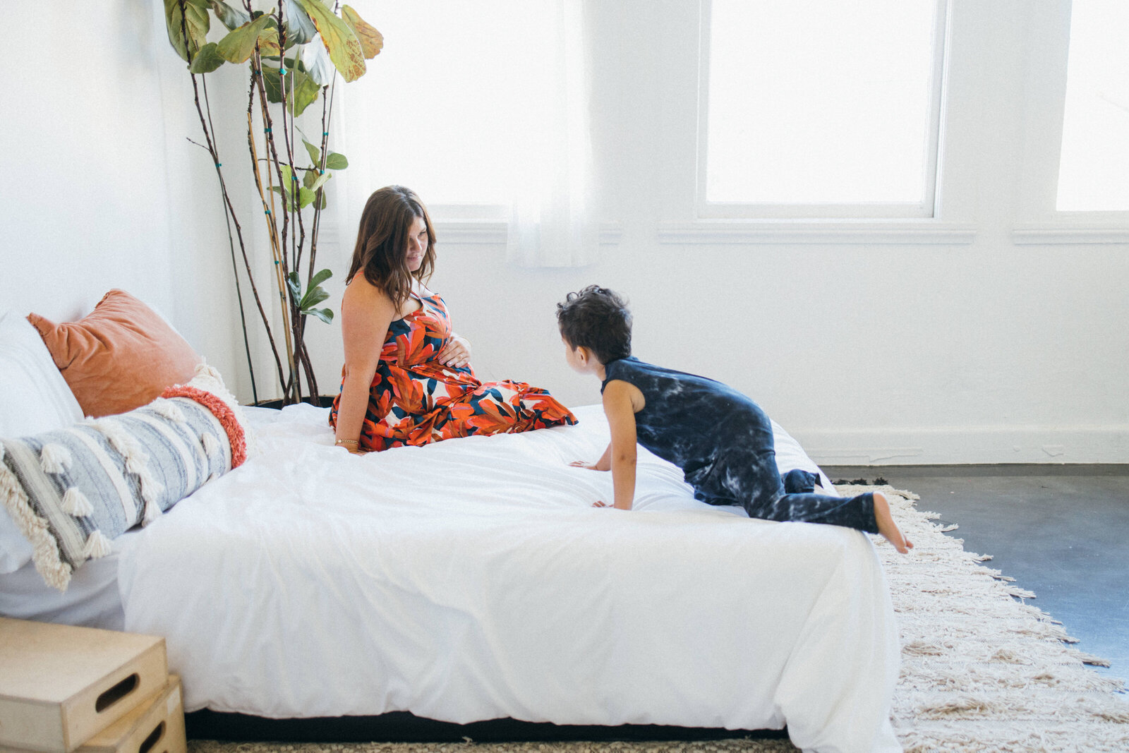 pregnant mom in red floral maxi dress sitting on the edge of a bed while her toddler in a blue romper crawls on the bed