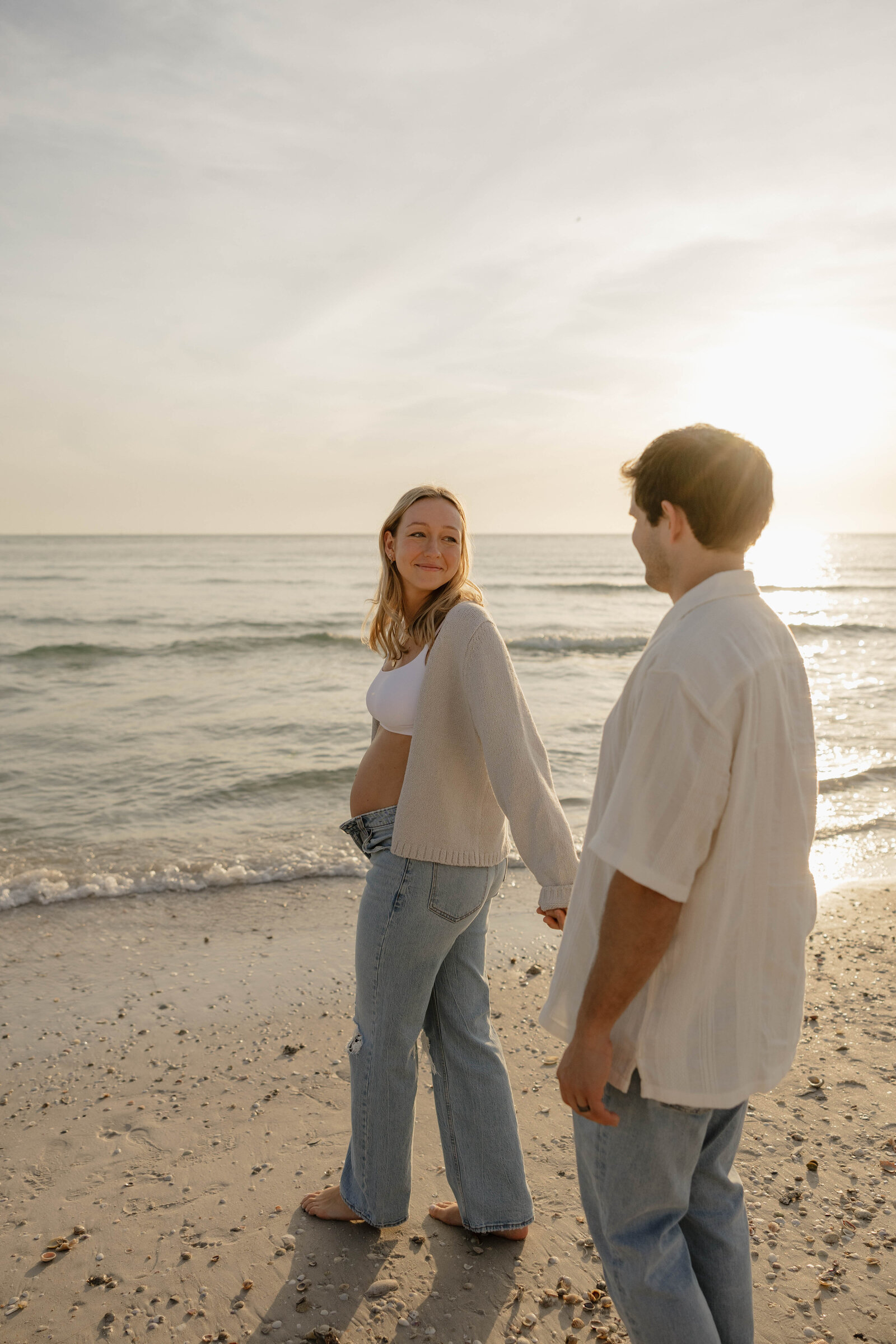 Couple holding hands and laughing during a photo shoot, surrounded by the water and soft sunlight.