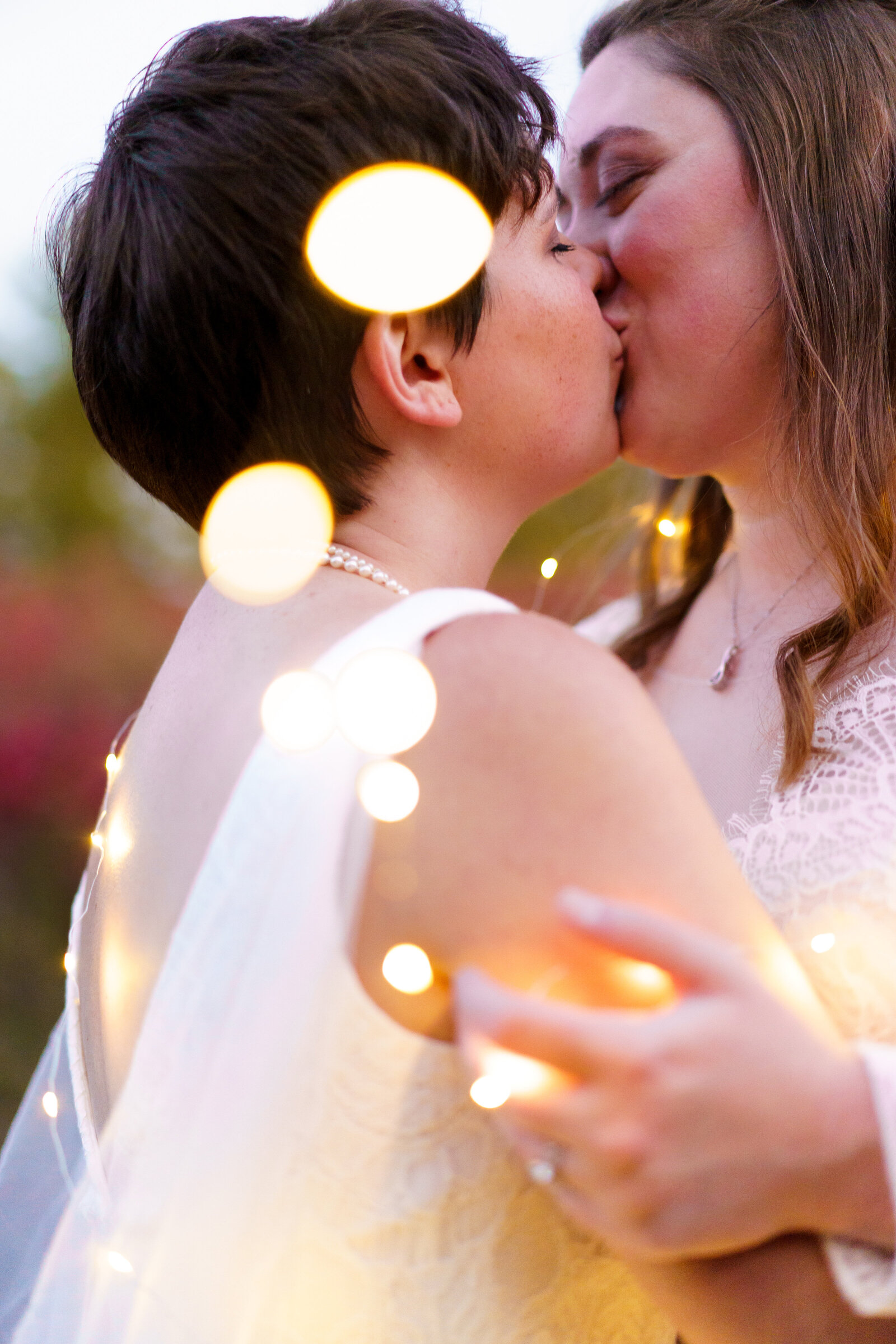 Two brides share a kiss while wrapped in fairy lights at sunset on their wedding day at Cedar Grove Lodging and Events in Hocking Hills.