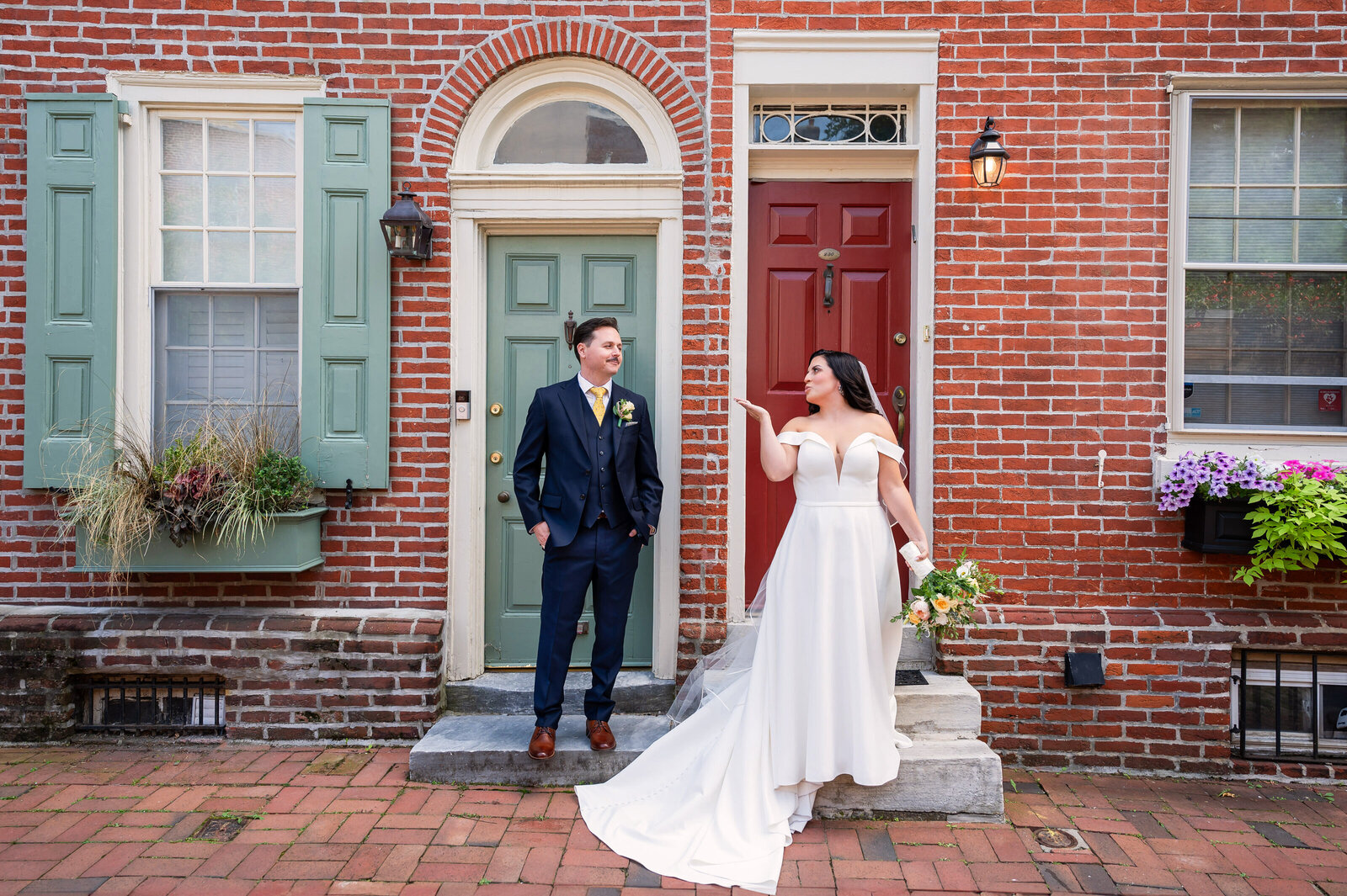 A bride and groom stand in front of a brick building with green and red doors. The bride wears a white dress and holds flowers, while the groom wears a dark suit. She playfully gestures toward him.