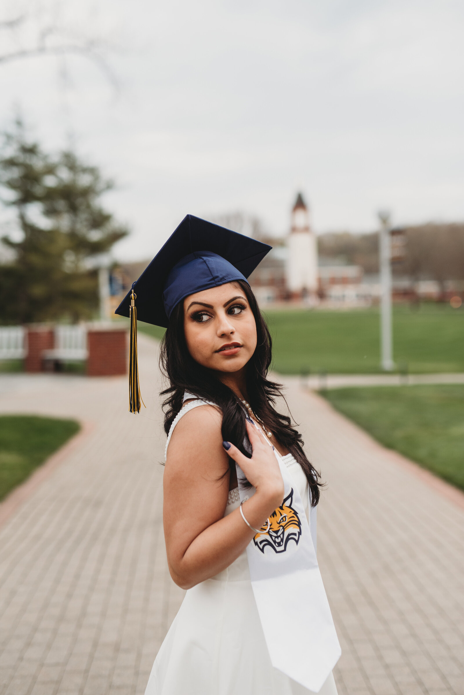 A Quinnipiac University graduate in a navy blue cap and white stole with the Bobcat logo poses on campus with the iconic clock tower in the background. Captured during a Connecticut college graduation photography session by Nicole Mele Photography, highlighting confidence and achievement.