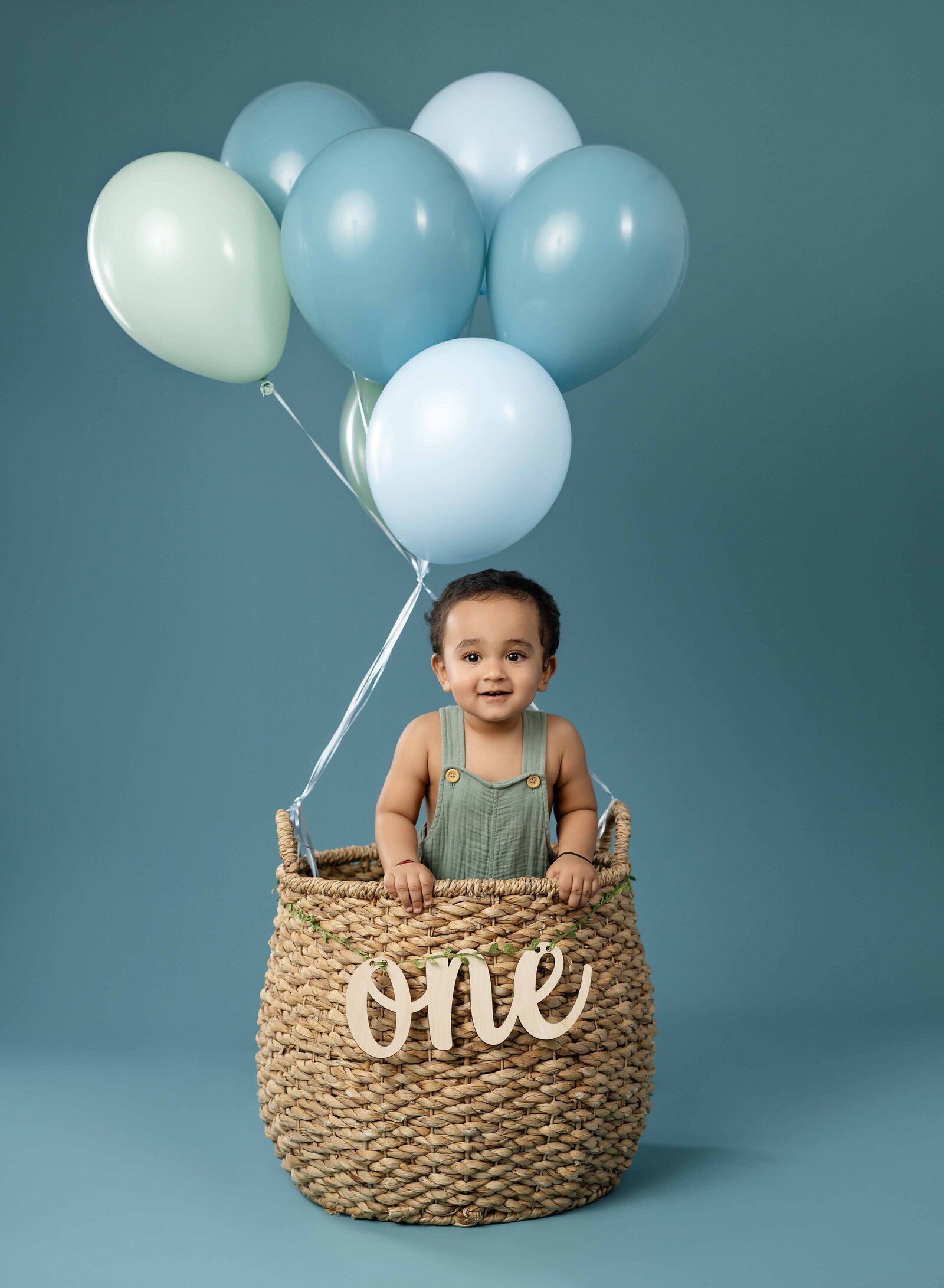 baby in birthday balloon basket with ONE letters in front and smiling at the camera.