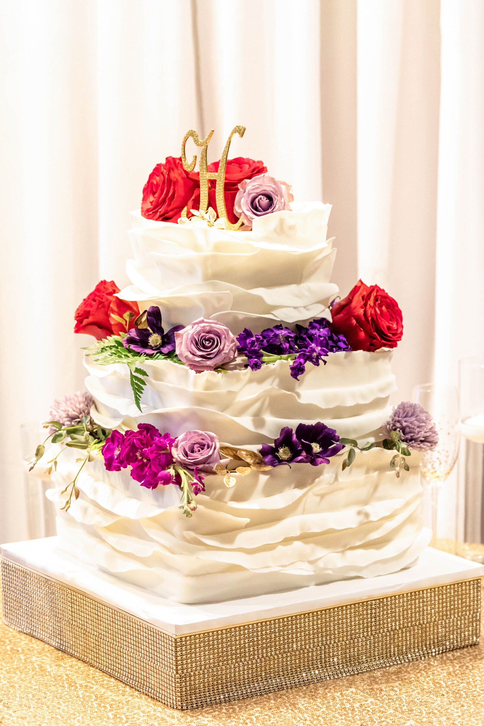 A stunning three-tiered wedding cake adorned with red, purple, and lavender flowers, topped with a golden "H" monogram, captured during a Ashleville, NC wedding, blending elegance and bold design.