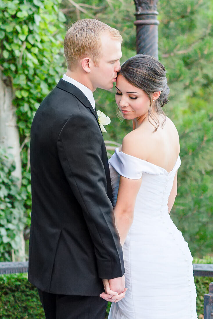A groom in a black tuxedo and a bride in an off the shoulder white wedding dress embrace under a pergola. Captured at the Sleepy Ridge Golf Course in Orem