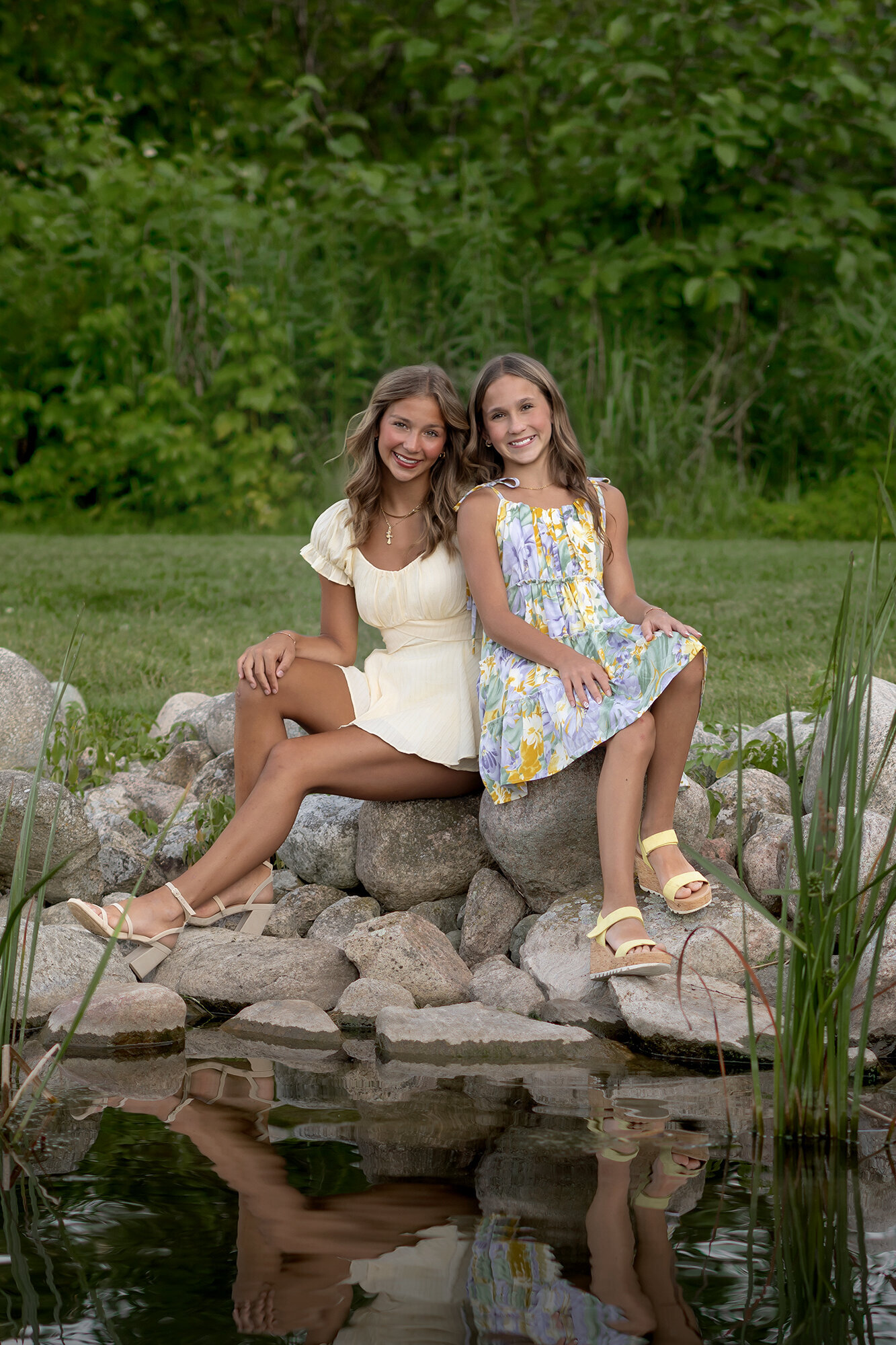 2 sisters lean against each other as they sit next to a pond