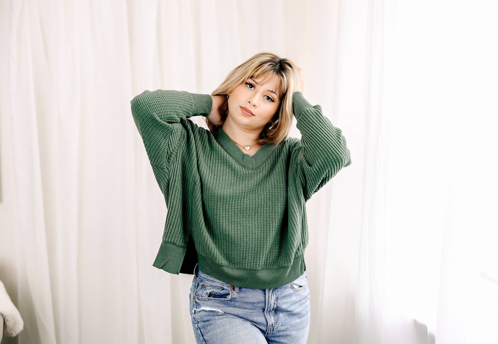 A person with shoulder-length blonde hair is standing with their hands behind their head, wearing a green ribbed sweater and light blue jeans. The background is a softly lit, light-colored curtain.