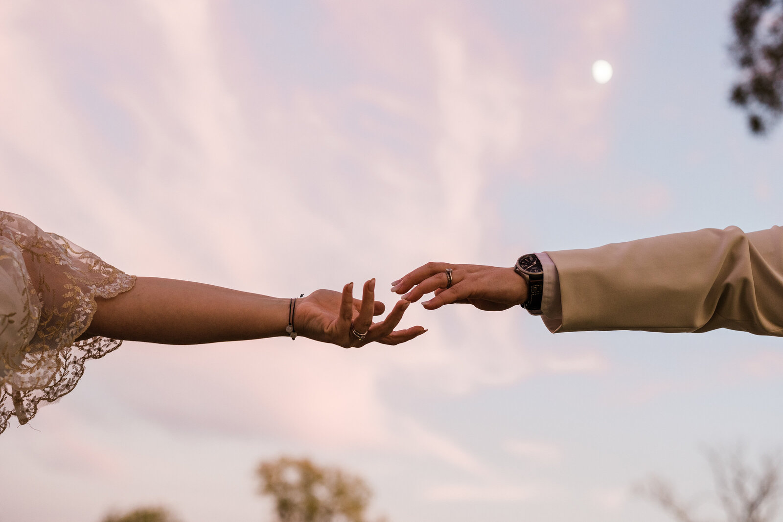 A creative wedding photo featuring two brides' hands reaching towards each other. The sunset sky is behind their hands, and it's pink and also features the moon.