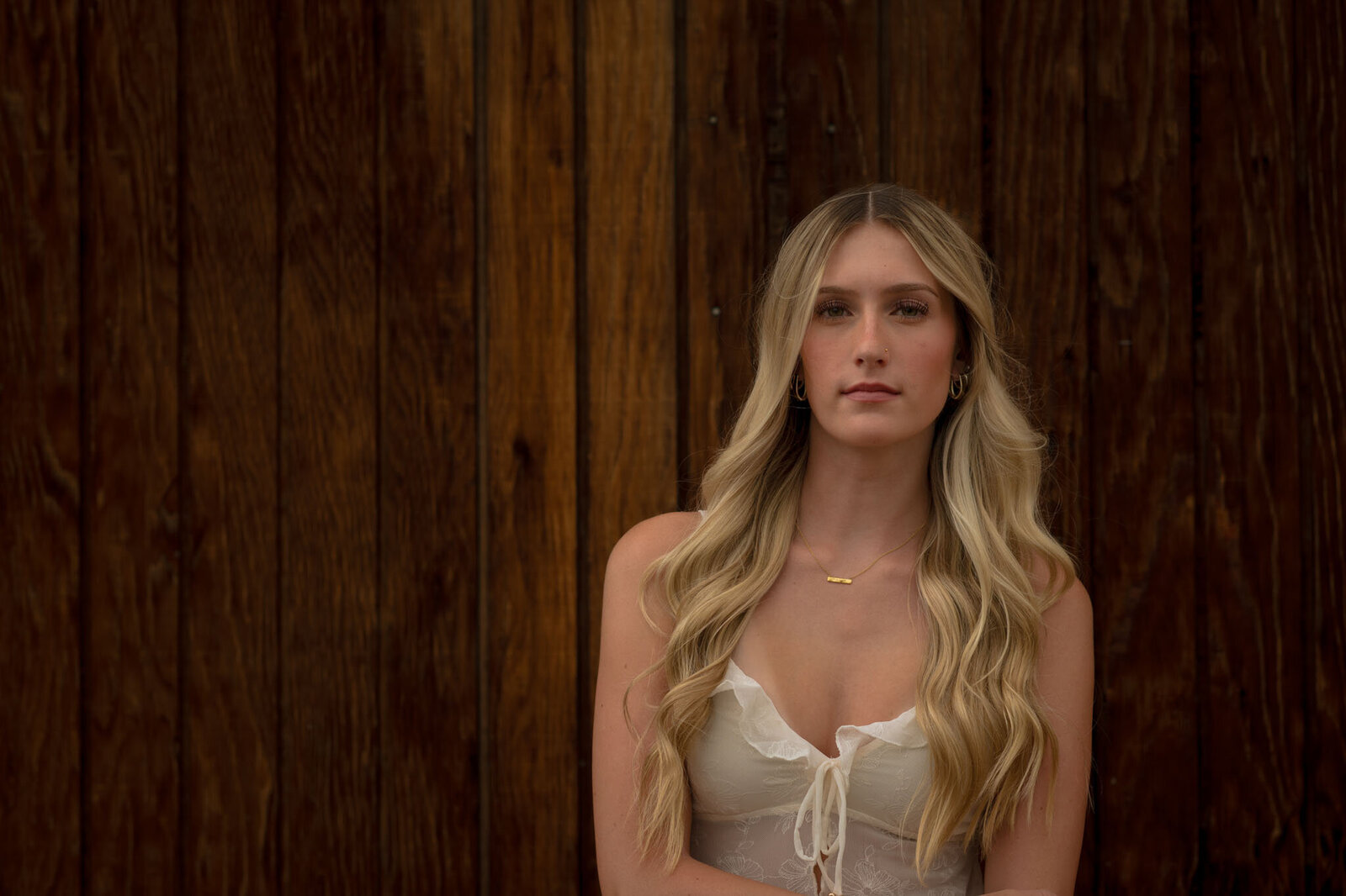 Teen girl with long blonde hair wearing a white top, standing against a rustic wooden wall during an outdoor senior photo session in Colorado.
